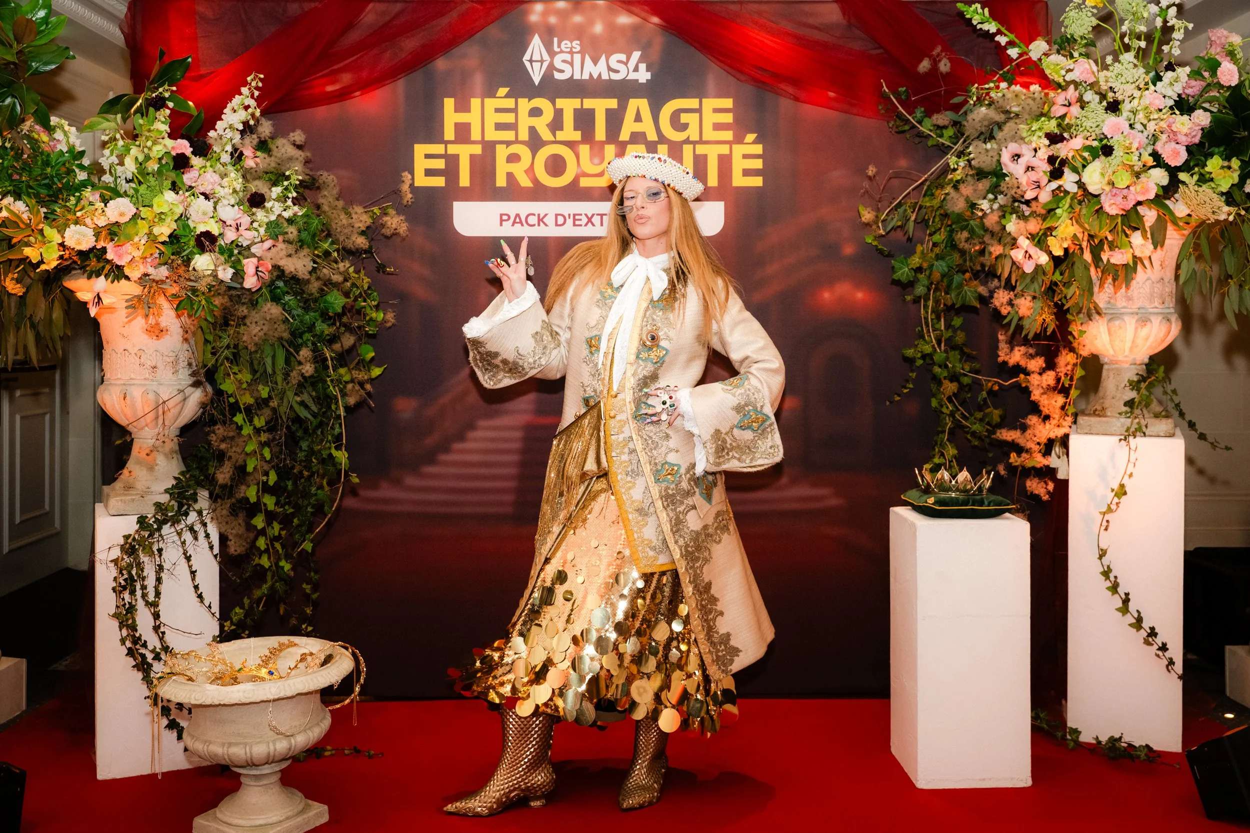 Une femme portant une robe longue en tissus dorés et décorée de sequins, avec un chapeau blanc orné de perles, pose devant un décor floral avec un panneau indiquant 'Les Sims 4: Héritage et Royauté, Pack d'Extensions'.
