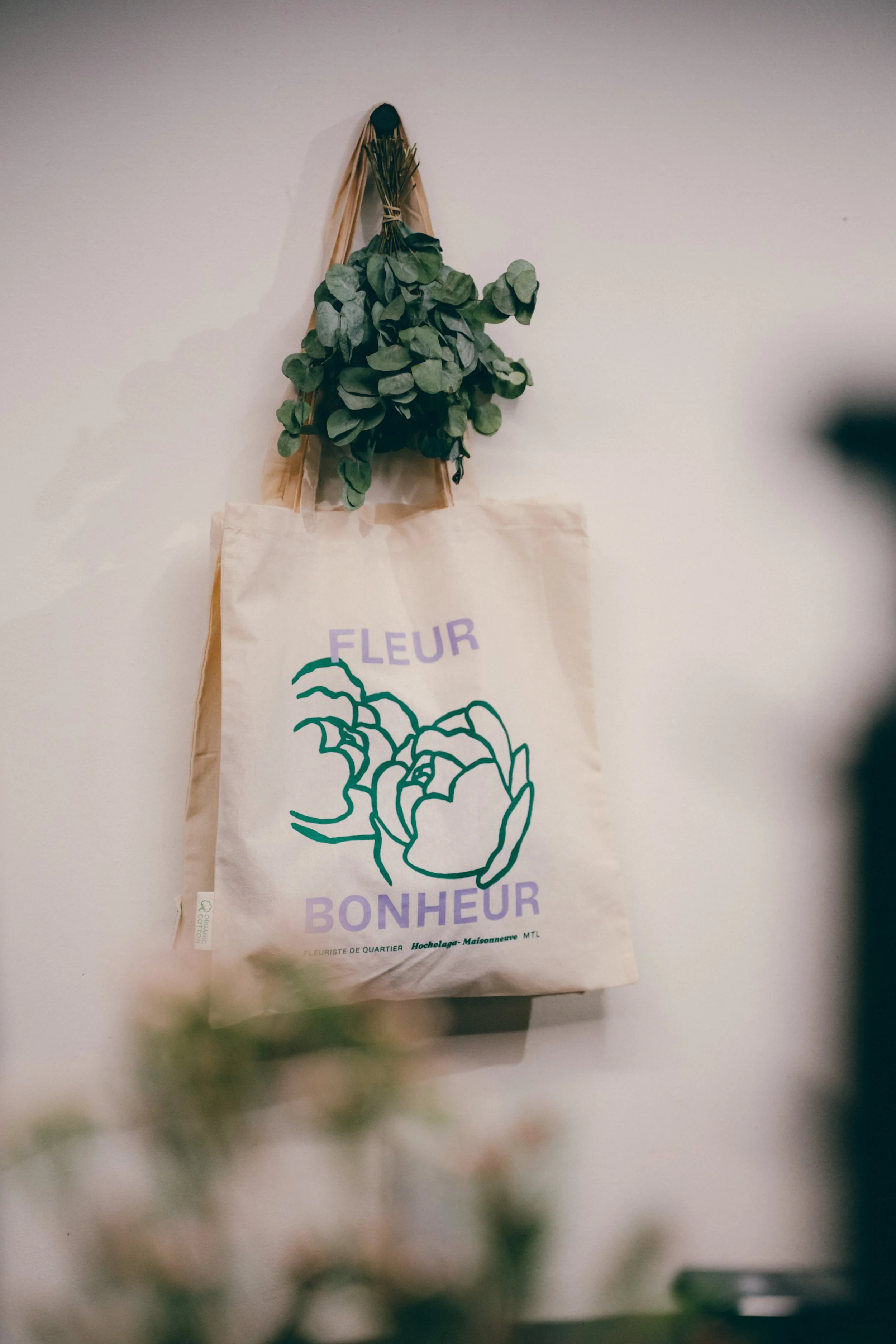 Un sac en papier avec une impression de dessin de fleur et le texte 'Fleur Bonheur', suspendu à un mur blanc avec un bouquet de feuilles vertes accrochées au-dessus.