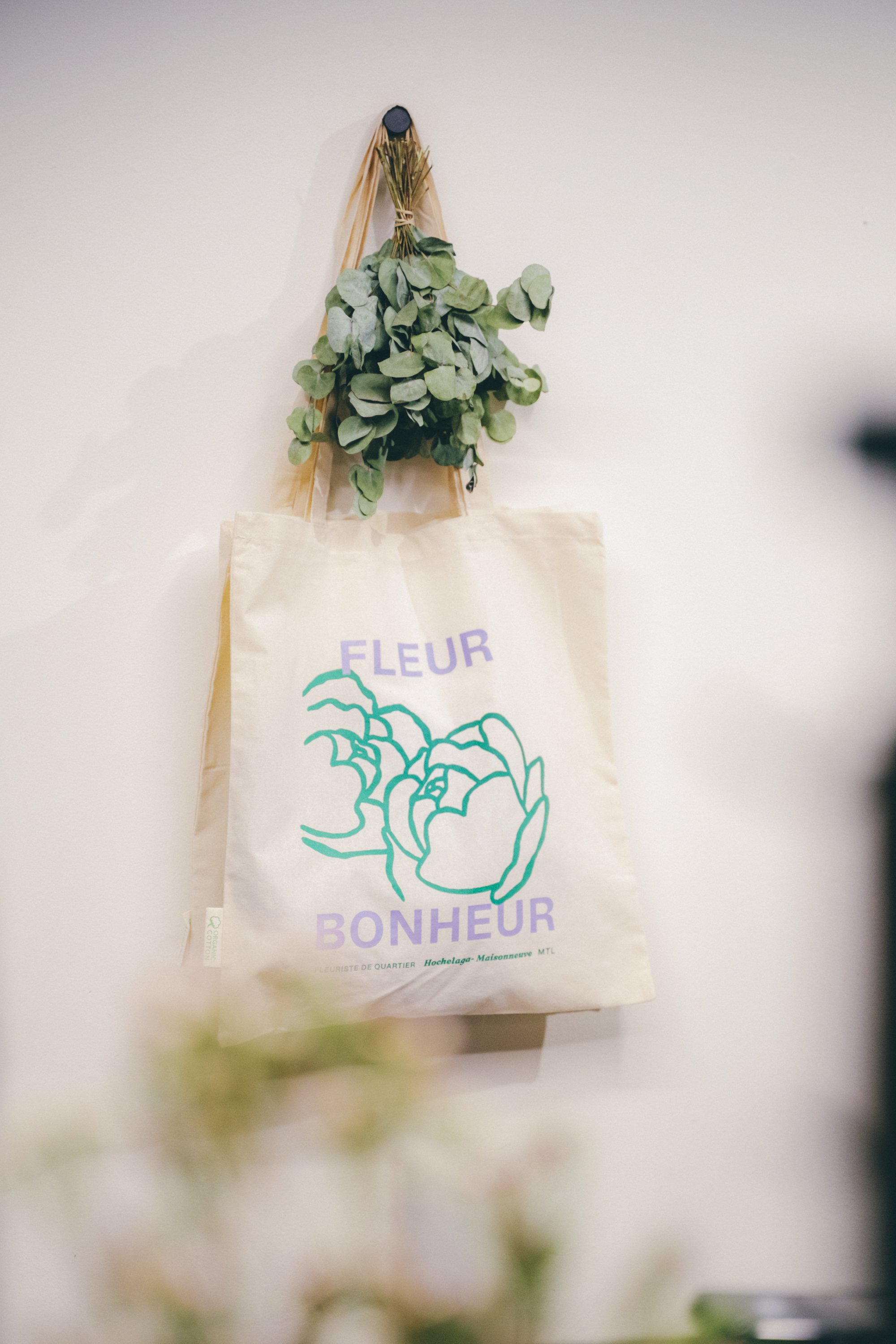 Un sac en papier avec une impression de dessin de fleur et le texte 'Fleur Bonheur', suspendu à un mur blanc avec un bouquet de feuilles vertes accrochées au-dessus.