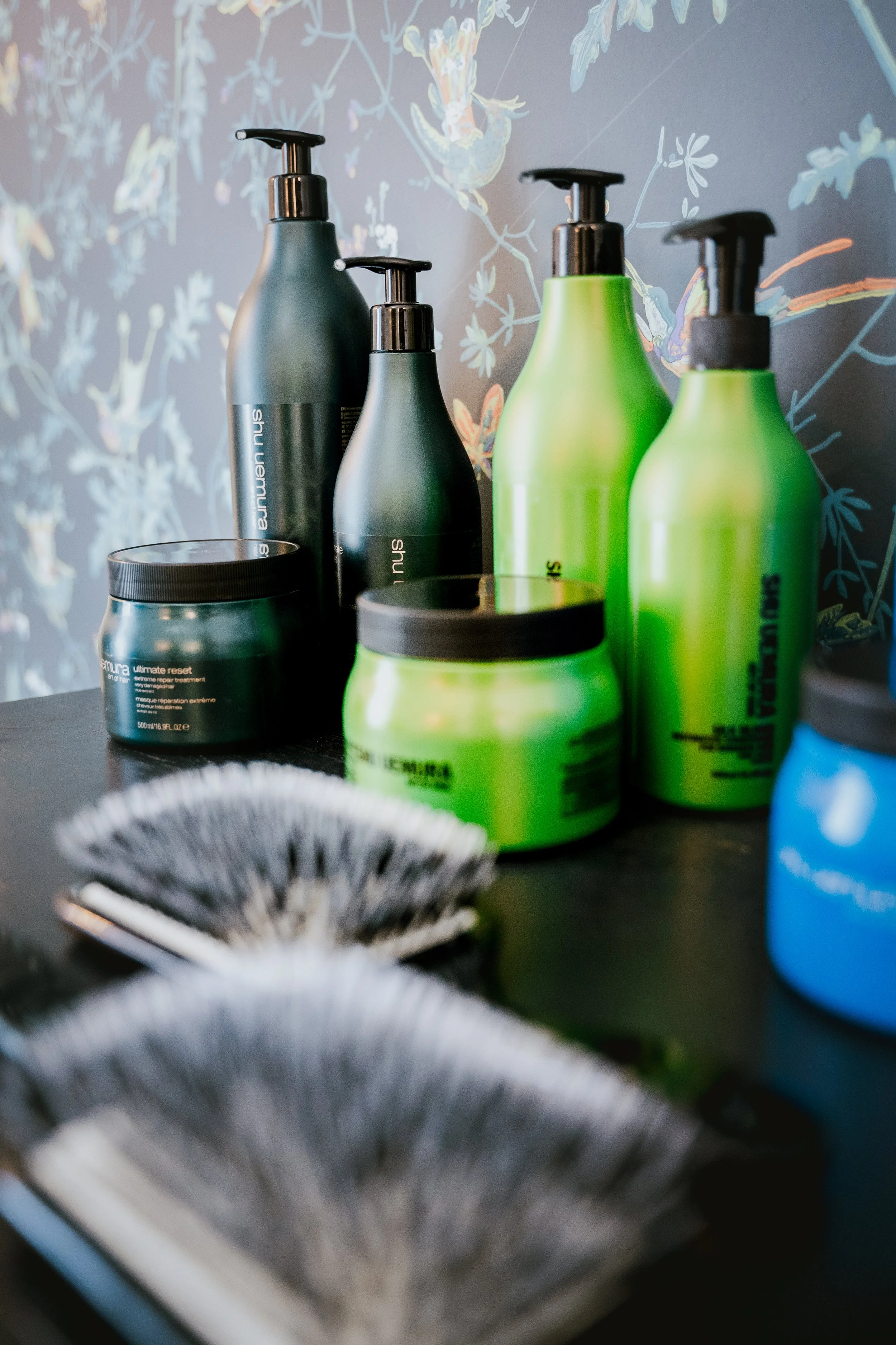 Une série de bouteilles de soins capillaires de différentes tailles en vert, gris et bleu sur une surface noire, avec un fond floral. Des pots de masque capillaire et deux brosses sont également visibles.