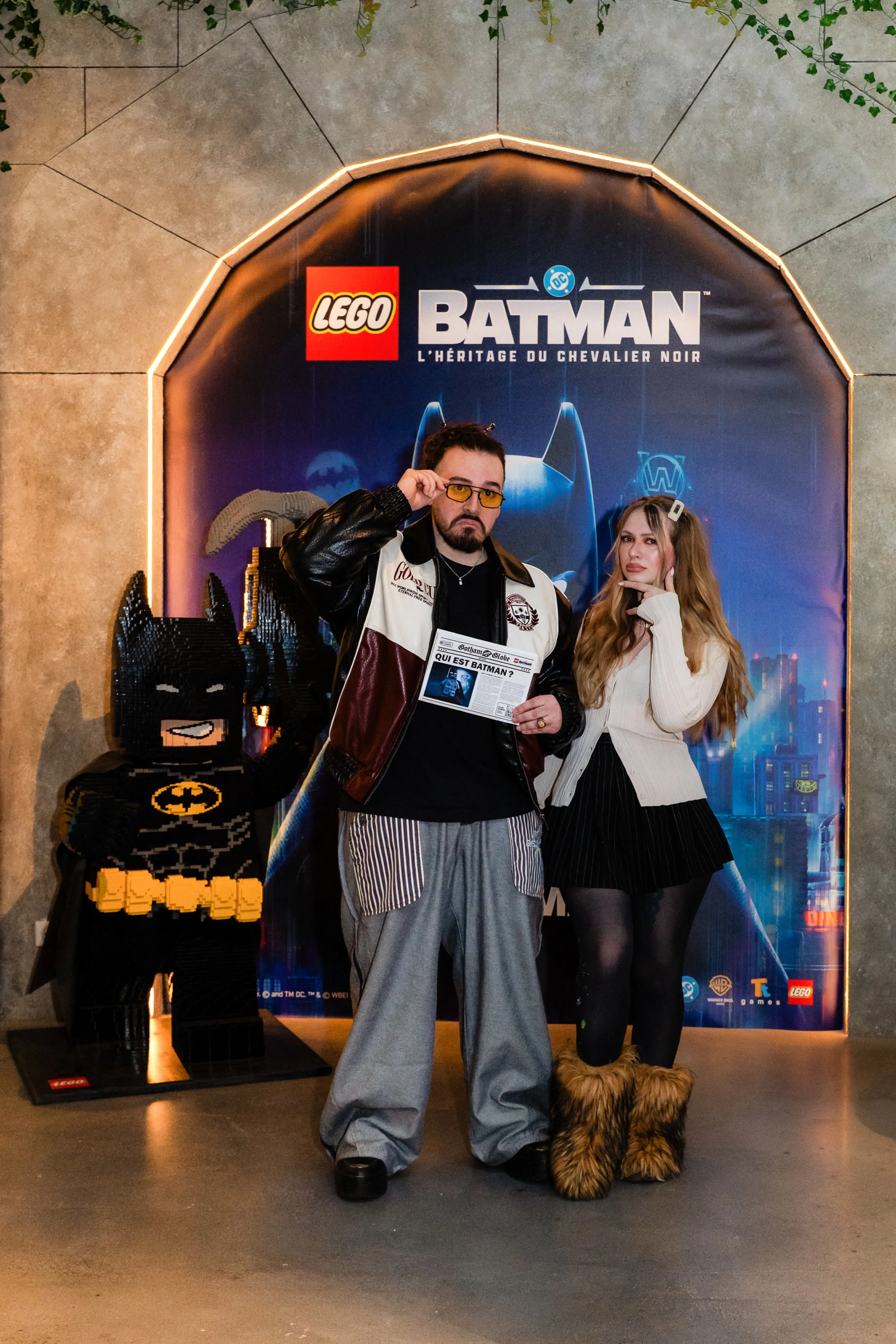 Deux personnes déguisées en personnages de Batman posent devant une affiche promotionnelle de LEGO Batman. La personne à gauche porte une veste en cuir et des lunettes jaunes, tenant un magazine, et est accompagnée d'un chien en costume de Batman ave