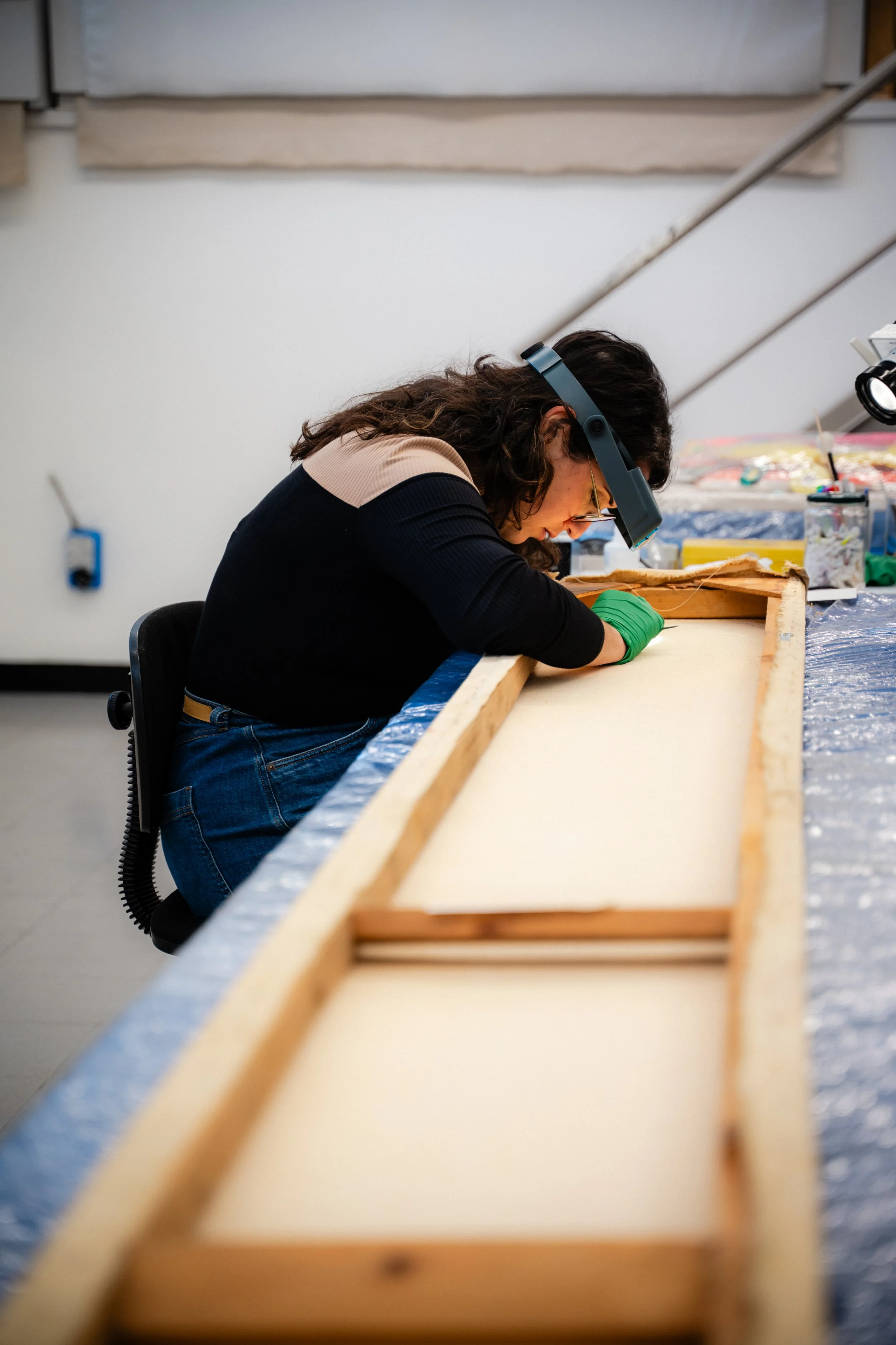 Une femme coiffée de lunette de protection et de gants verts travaille sur une grande surface en atelier, manipulant un grand tableau sur une table de travail.