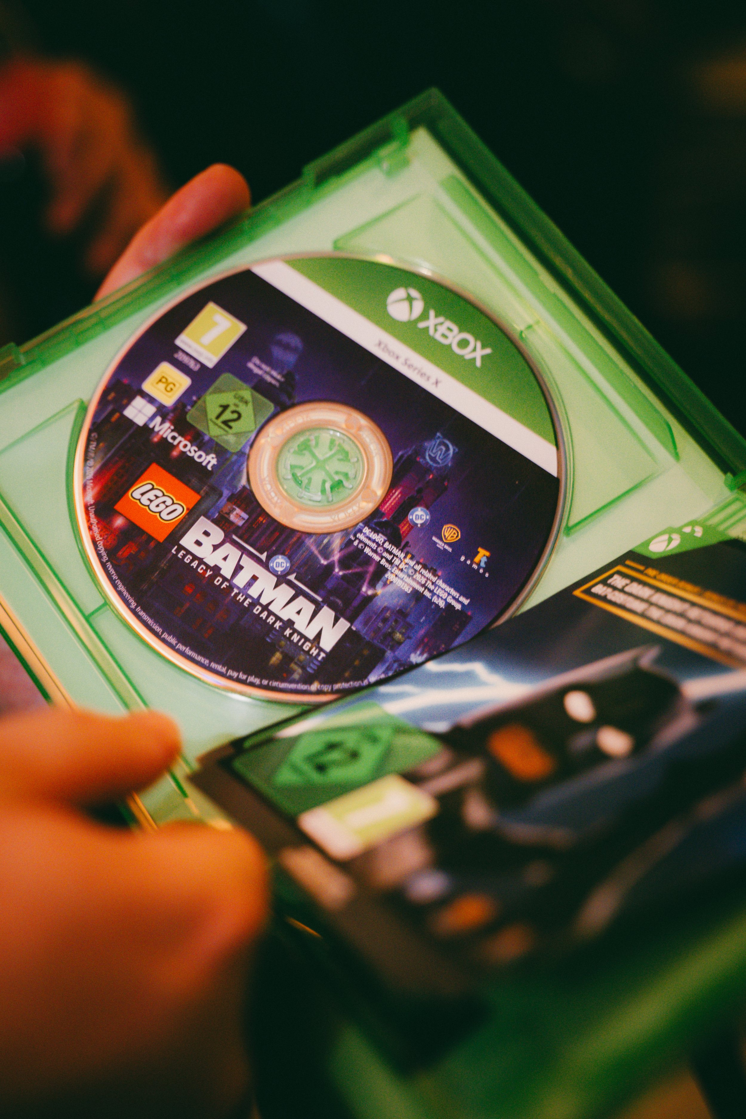 Une main tenant une boîte en plastique vert contenant un CD du jeu vidéo « Battlefield : Legacy of the Dark Knight » pour la console Xbox. Le CD affiche le logo Xbox, le titre du jeu, et une illustration de Batman et le logo DC.