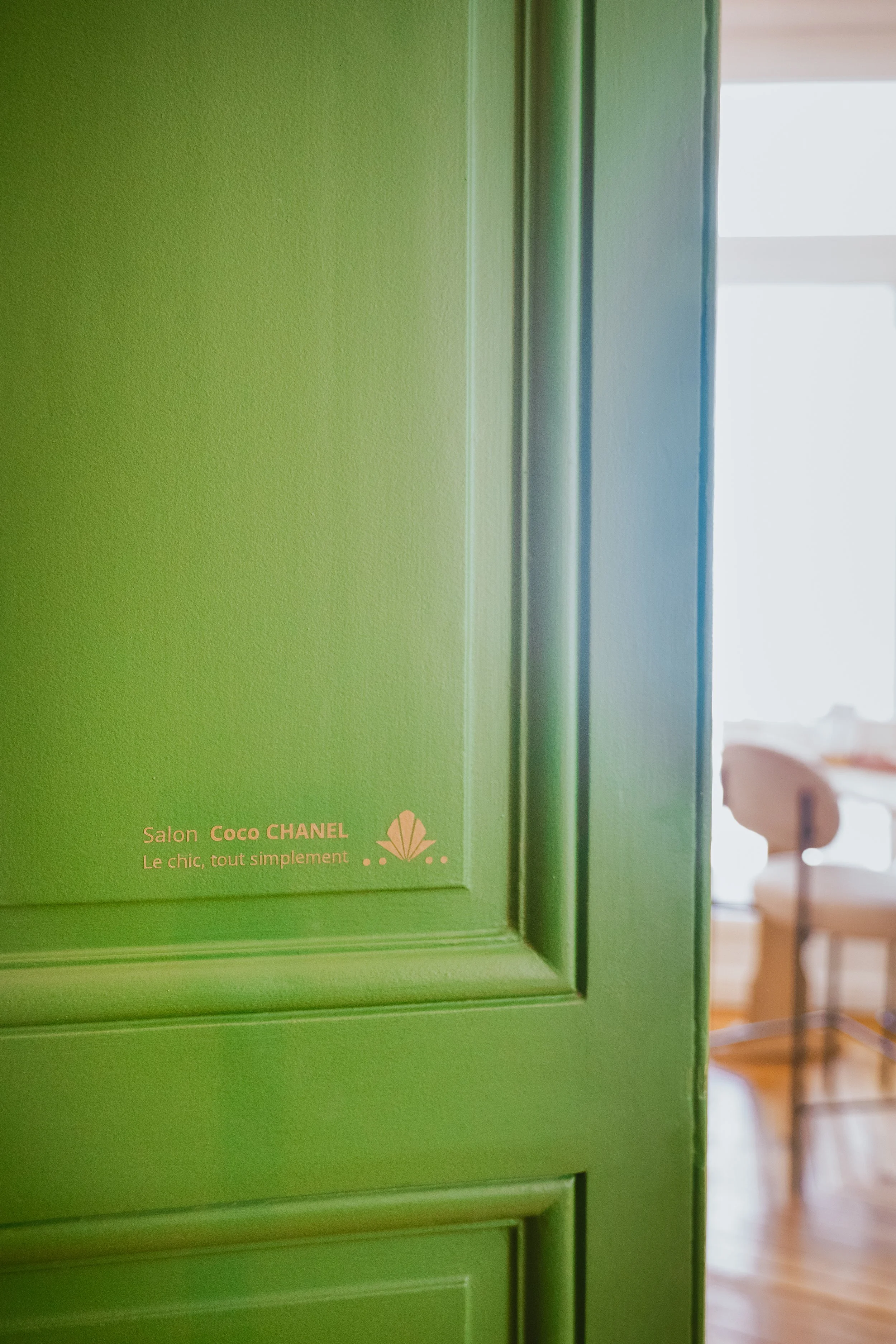Une porte verte, avec en bas à gauche un texte en français "Salon Coco CHANEL, Le chic, tout simplement". En arrière-plan, une pièce avec un fauteuil en bois, un tapis et un sol en bois visible à droite.