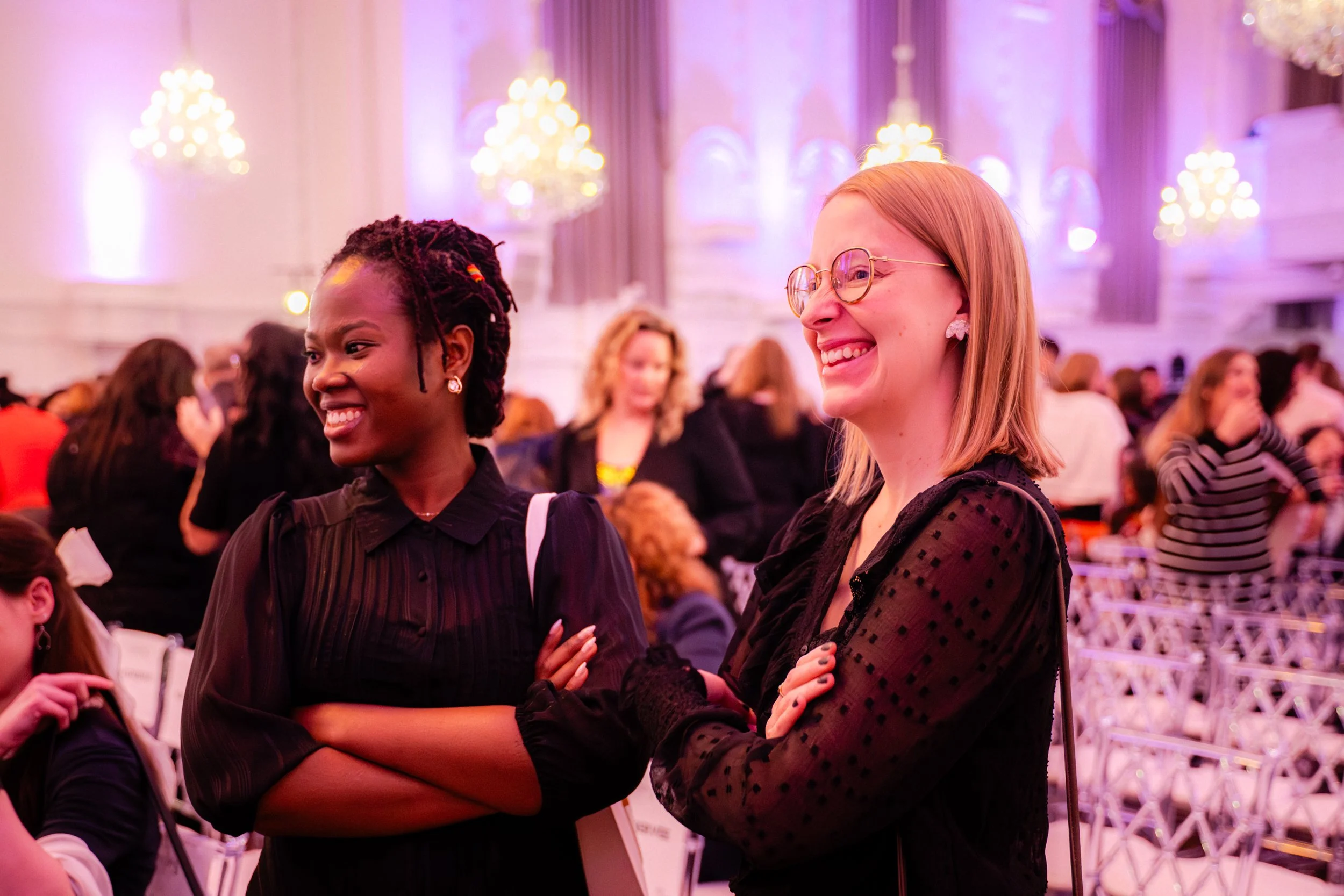 Deux femmes souriantes dans une salle élégante, probablement lors d'un événement ou d'une réception, avec des chandeliers lumineux en arrière-plan.
