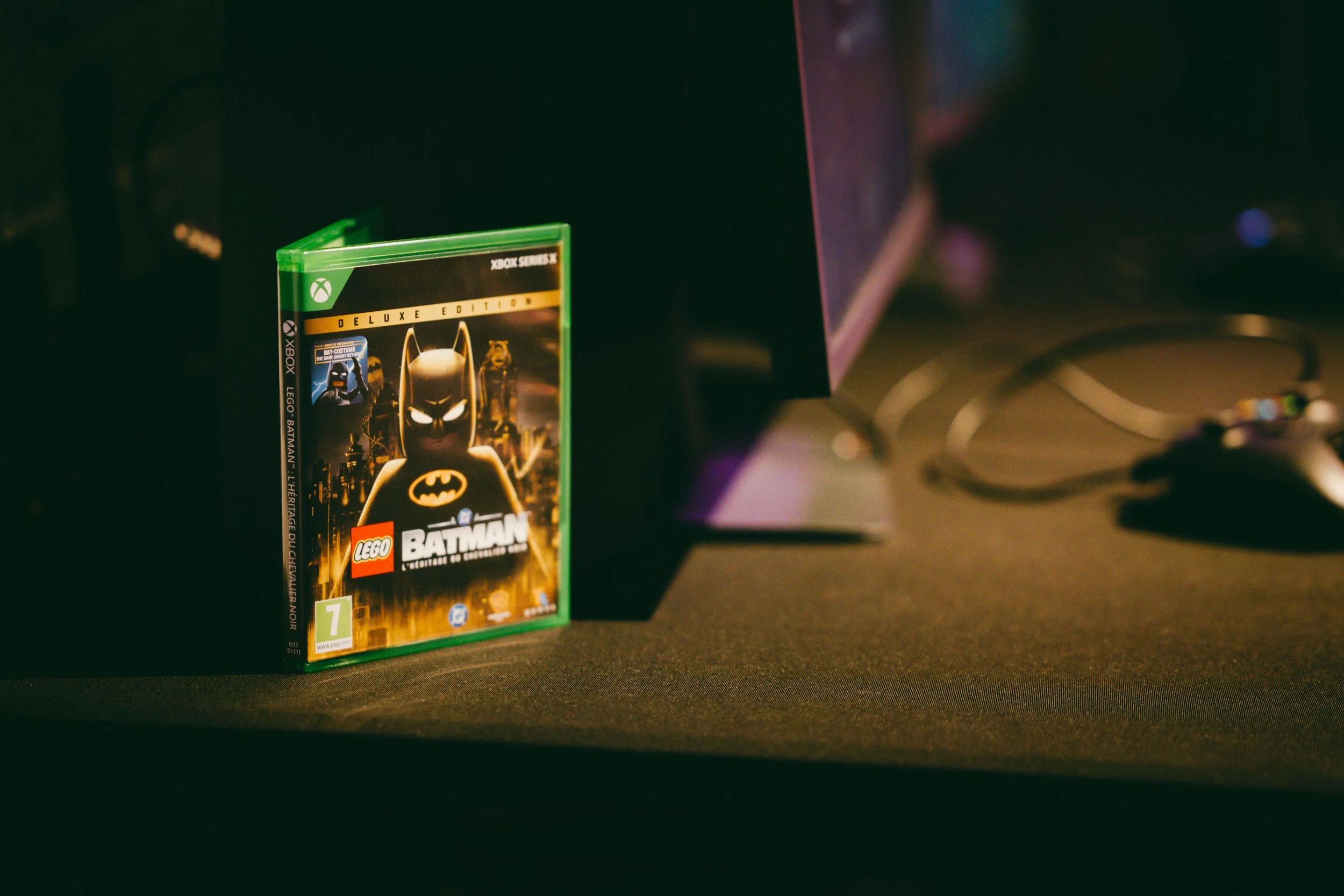 Étui de jeu vidéo Lego Batman 3 sur une surface noire, avec un fond sombre et un écran d'ordinateur partiellement visible sur le côté droit.