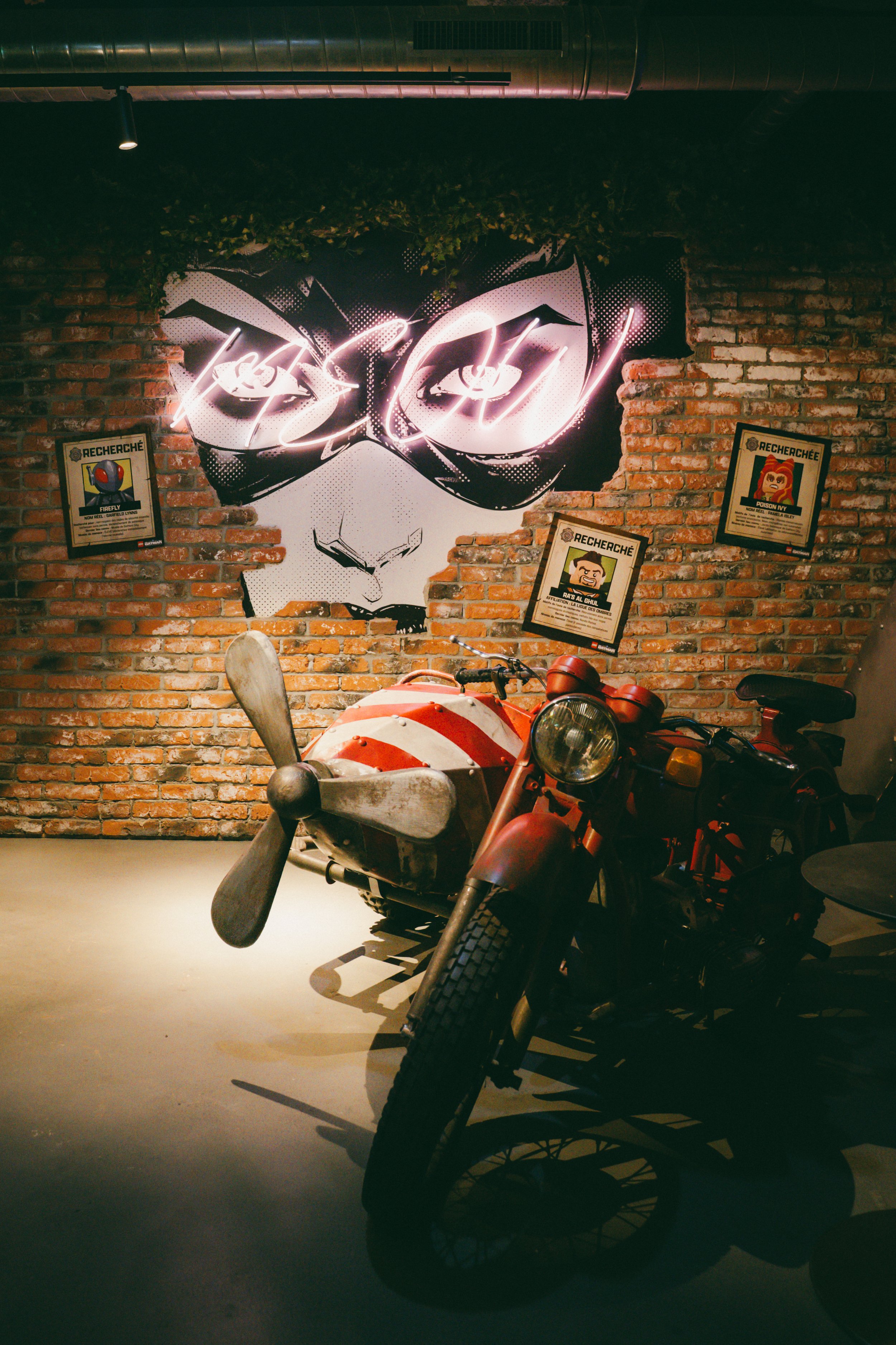 Une moto vintage dans un environnement intérieur avec mur en briques, décoré d'une œuvre d'art représentant un visage avec des lunettes de soleil. Trois affiches de recherche sont fixées au mur à gauche et à droite de l'œuvre.