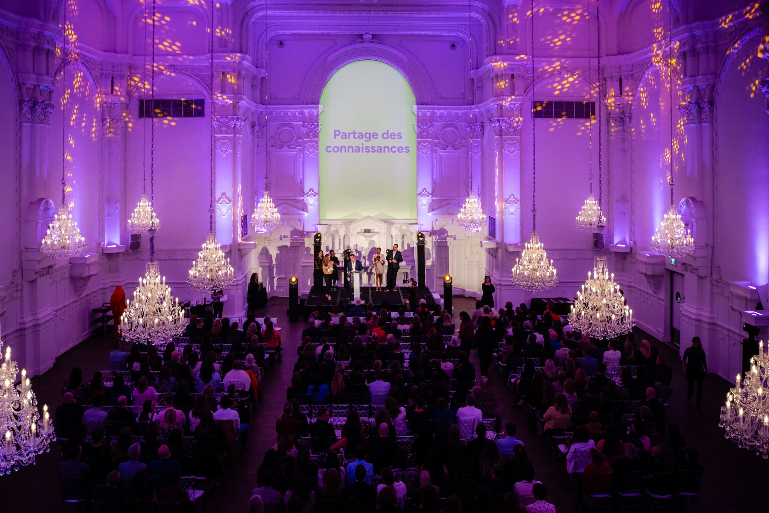 Une grande salle ornée de lustres, avec un groupe de personnes sur scène et un public assis. Le mur au fond affiche le message 'Partage des connaissances' en violet sur un écran lumineux vert.