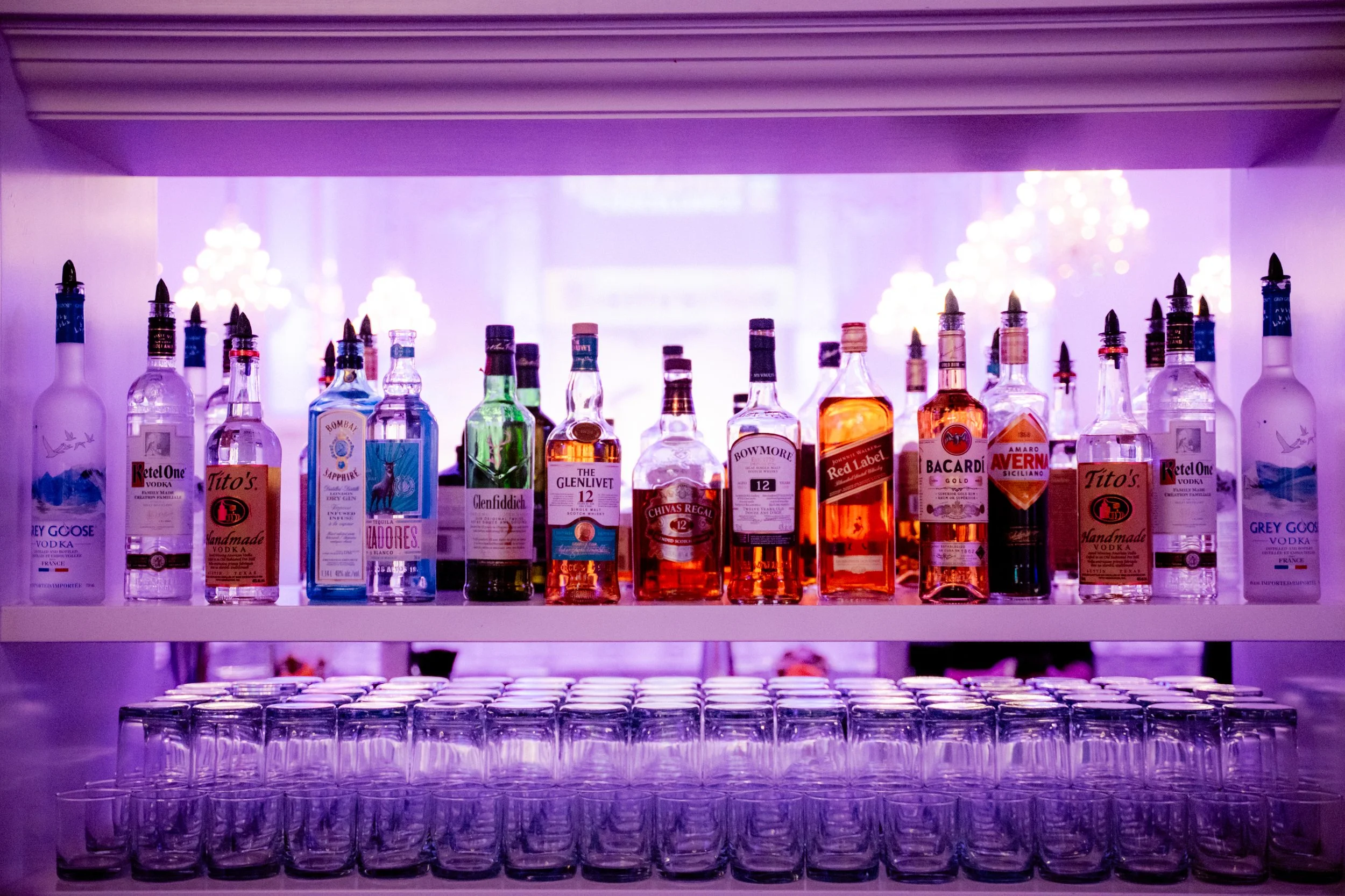 Une étagère de bouteilles d'alcool colorées sous un éclairage violet avec des verres empilés en dessous.