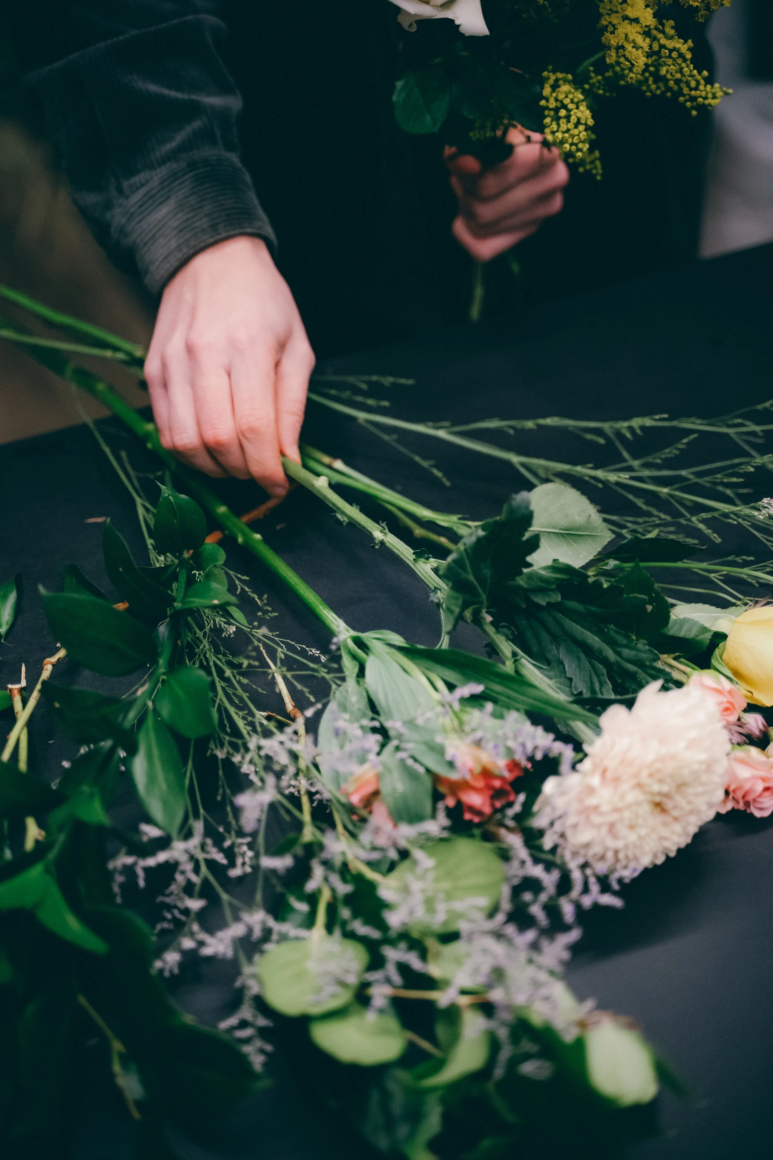 Mains masculines assemblant un bouquet de fleurs colorées comprenant des roses, des hortensias, et diverses plantes vertes sur une surface sombre.