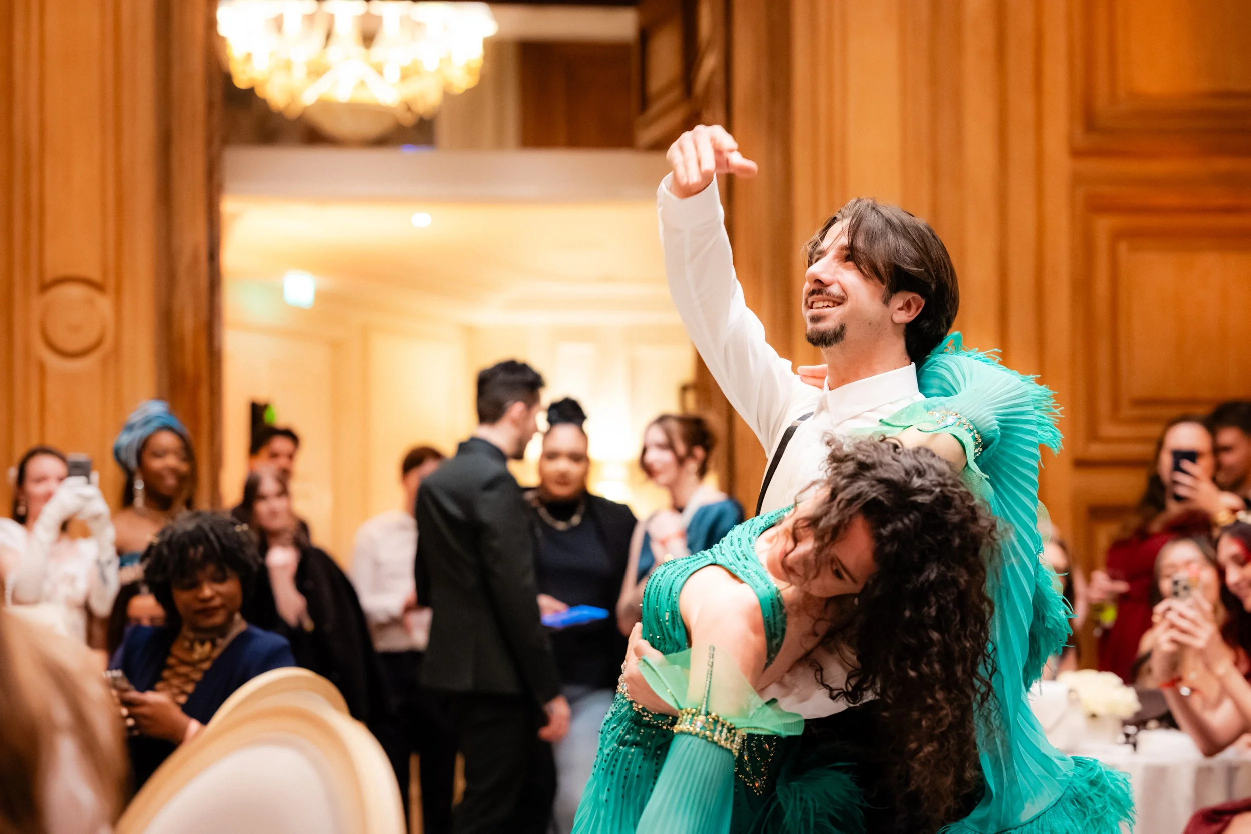 Une personne ou une danseuse danse avec une autre personne lors d'une célébration ou d'un événement social, entourée de spectateurs souriants dans une salle ornée de bois et d'un lustre chandelier.