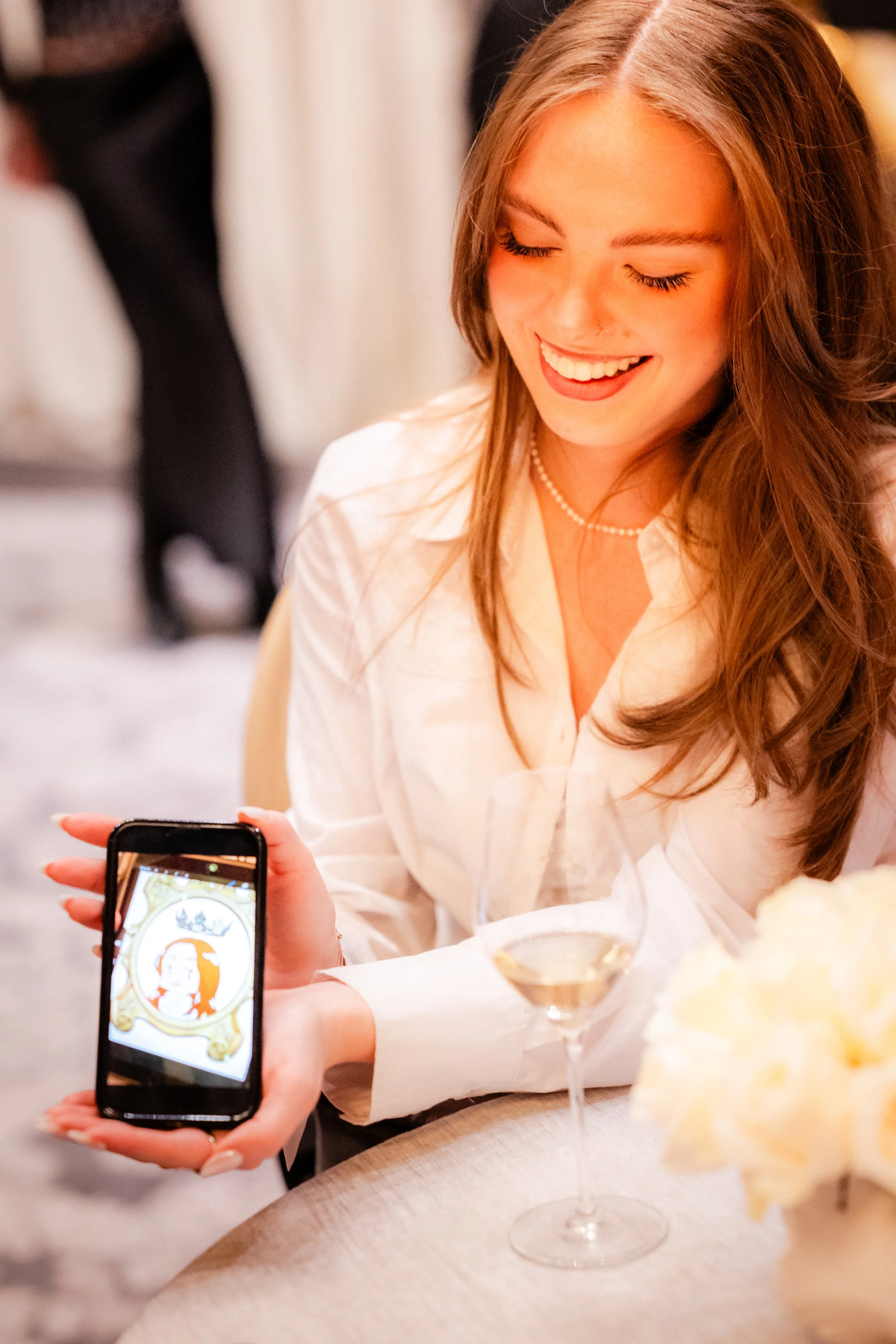 Une femme souriante portant une chemise blanche regarde un téléphone portable, dans un cadre lors d'une occasion sociale, avec un verre à cocktail devant elle et des détails dans l'arrière-plan.