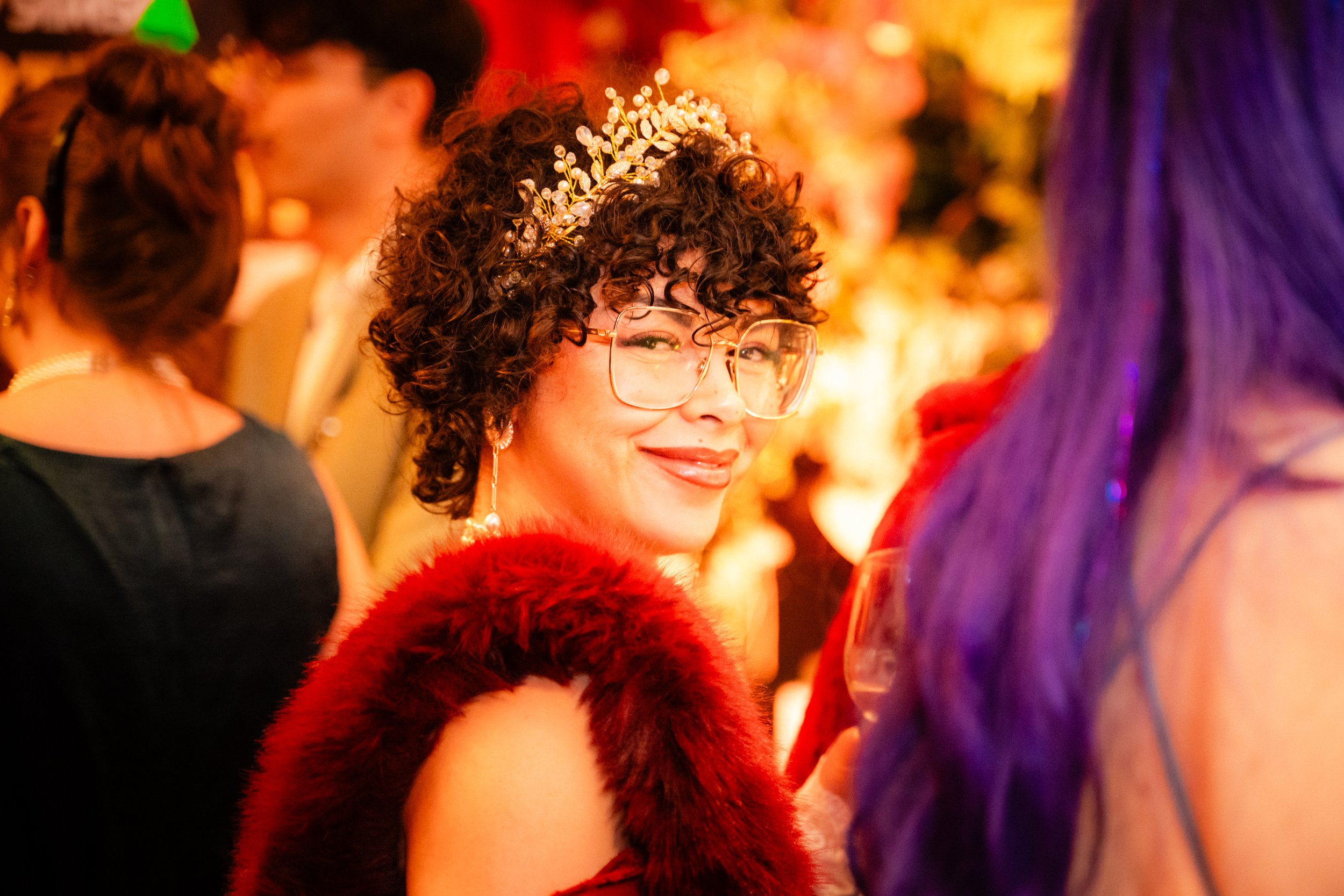 Femme souriante portant des lunettes, un serre-tête en perles, une robe en velours rouge et une cape en fourrure rouge, entourée de personnes à un événement festif.