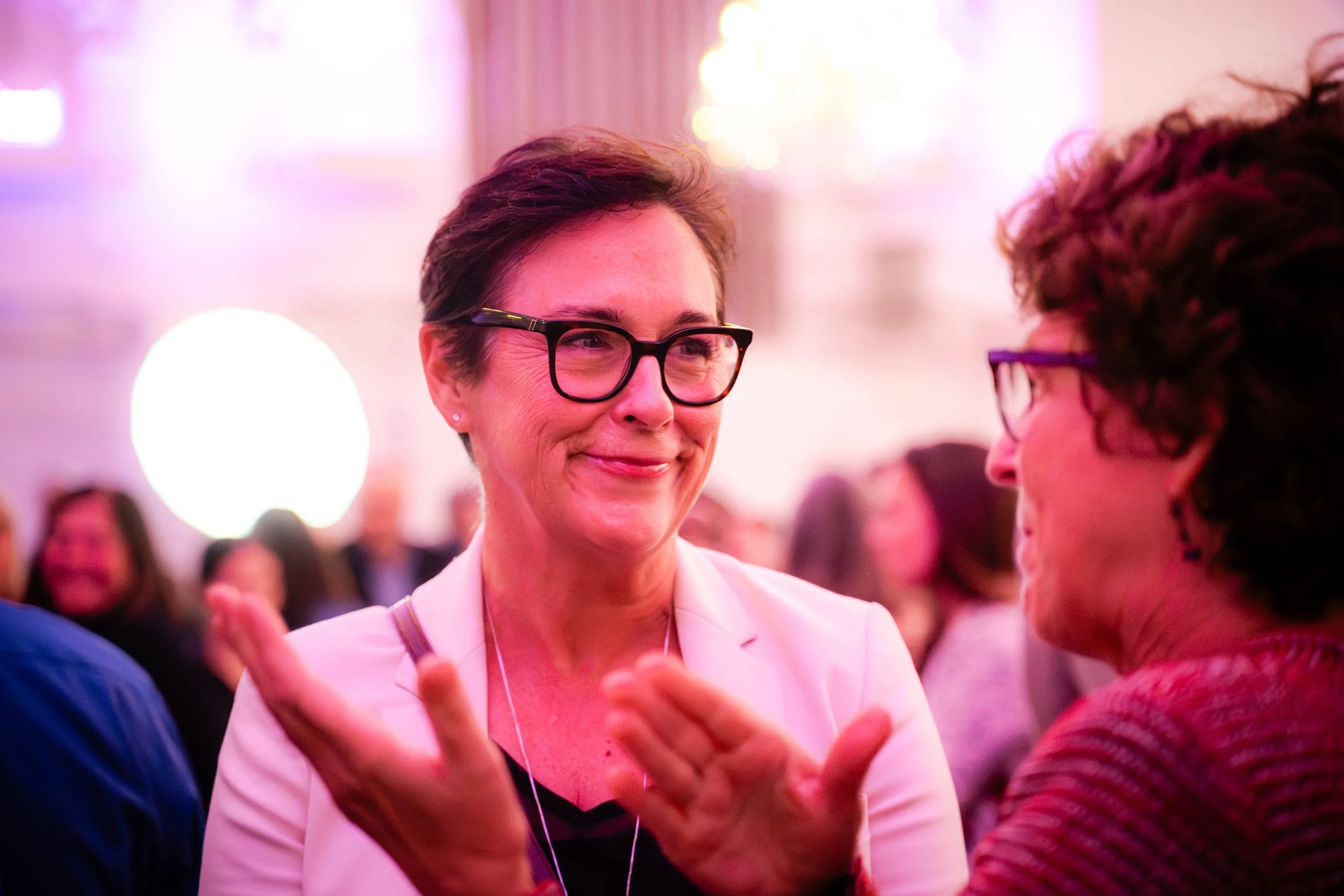 Une femme souriante porte des lunettes noires et discute avec une autre personne lors d'une soirée chaleureuse et colorée.