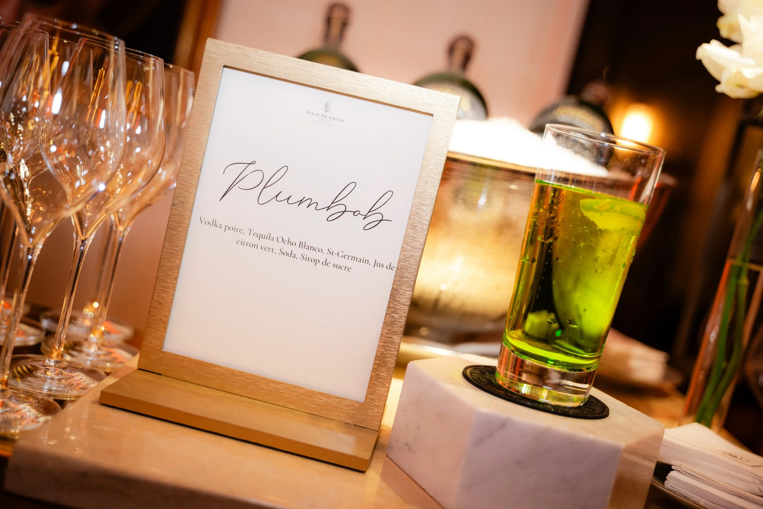 Verres à cocktail alignés avec un panneau indiquant une boisson nommée 'Plumbob' et la liste de ses ingrédients, tels que vodka poire, tequila, citron vert, soda, sirop de sucre, dans un environnement de bar ou de réception.
