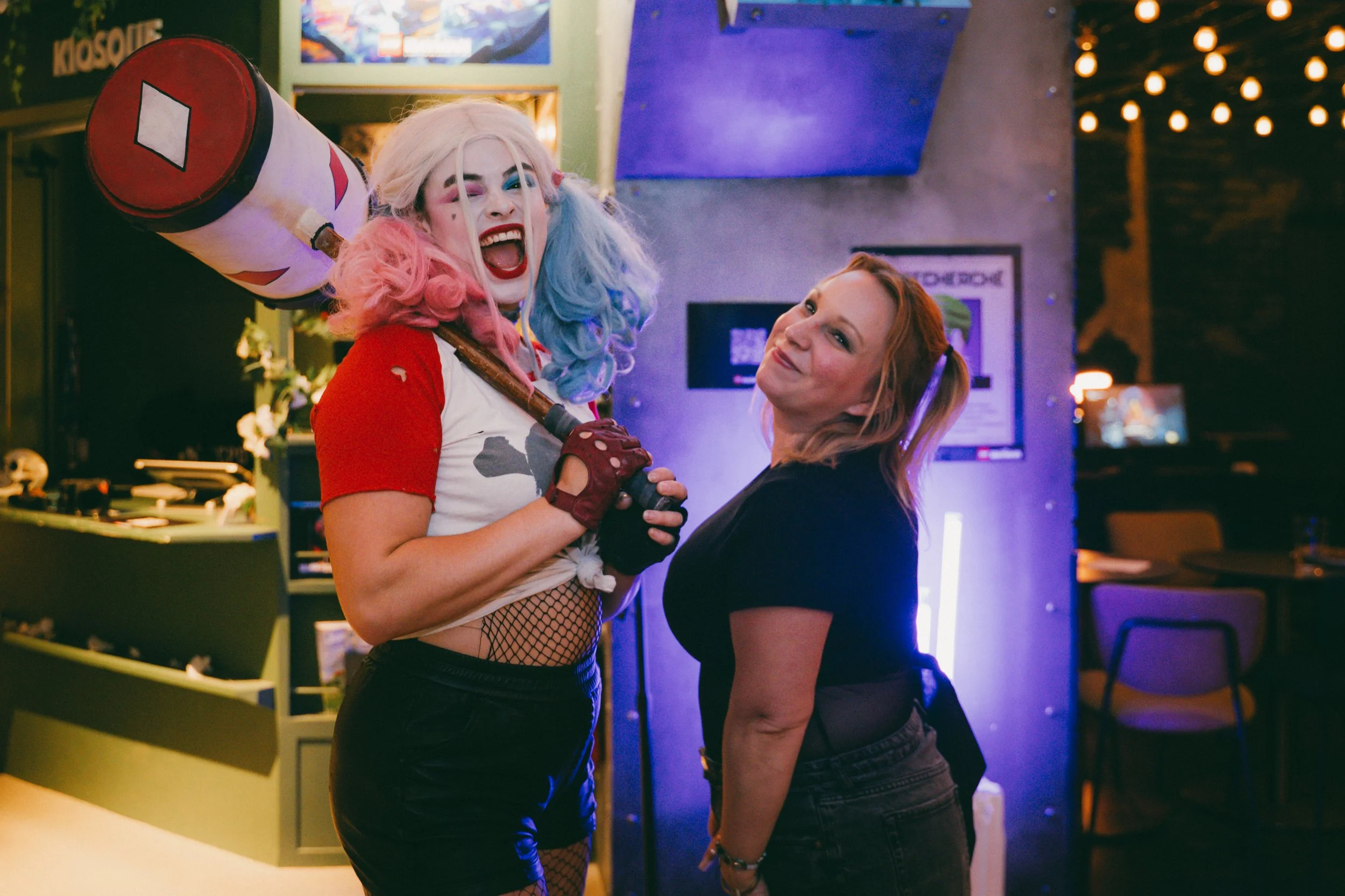 Deux femmes dans un lieu de divertissement, l'une portant une perruque rose et bleue, un maquillage coloré, et un t-shirt déchiré, brandissant une batte de baseball. L'autre, avec des cheveux roux, porte un t-shirt noir. Elles semblent partager un mo