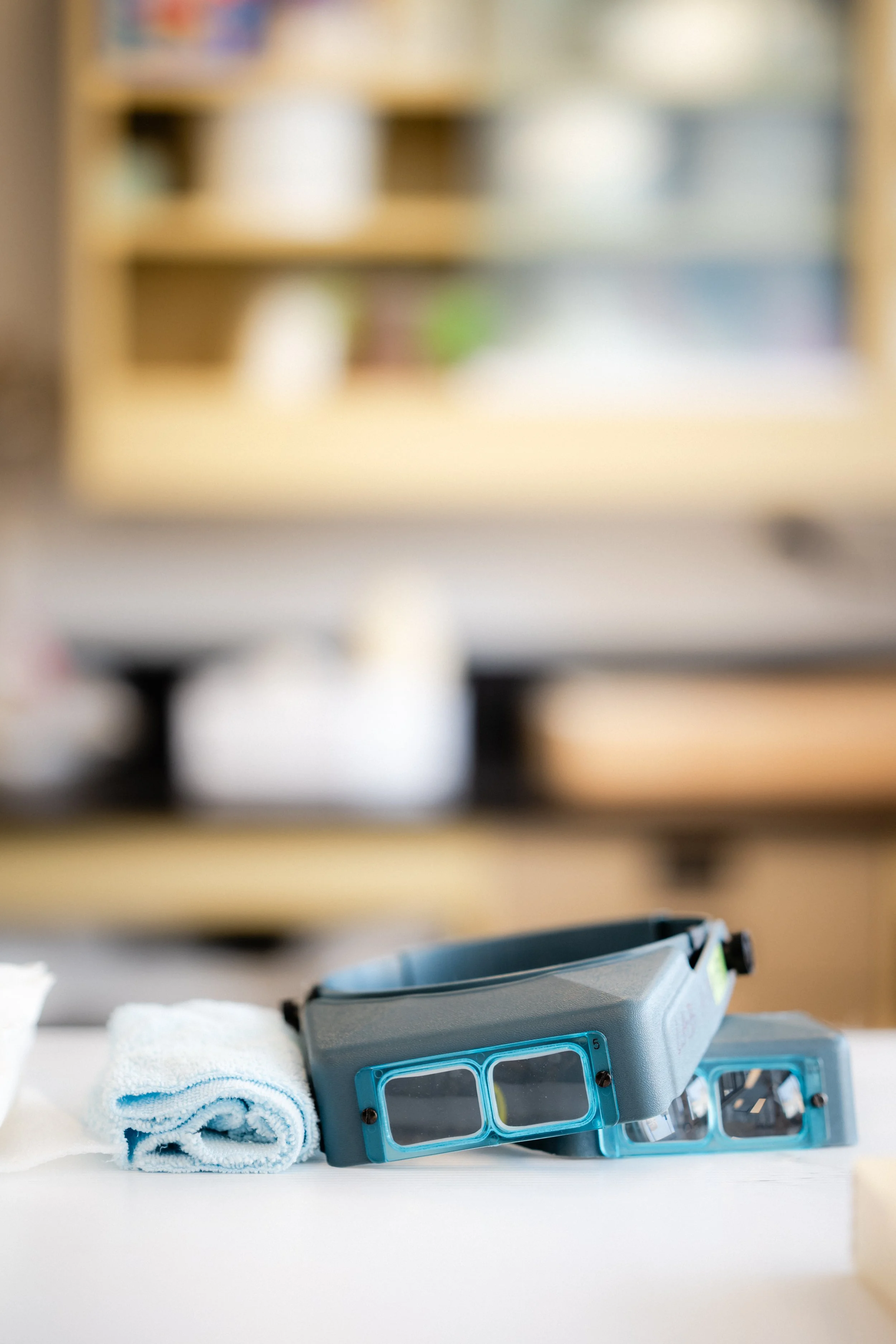 Une boîte de rangement en plastique avec des compartiments transparent, une serviette blanche, une serviette bleue, et un appareil de nettoyage ou de réparation sur une surface blanche. En arrière-plan, une étagère en bois avec des livres ou des obje
