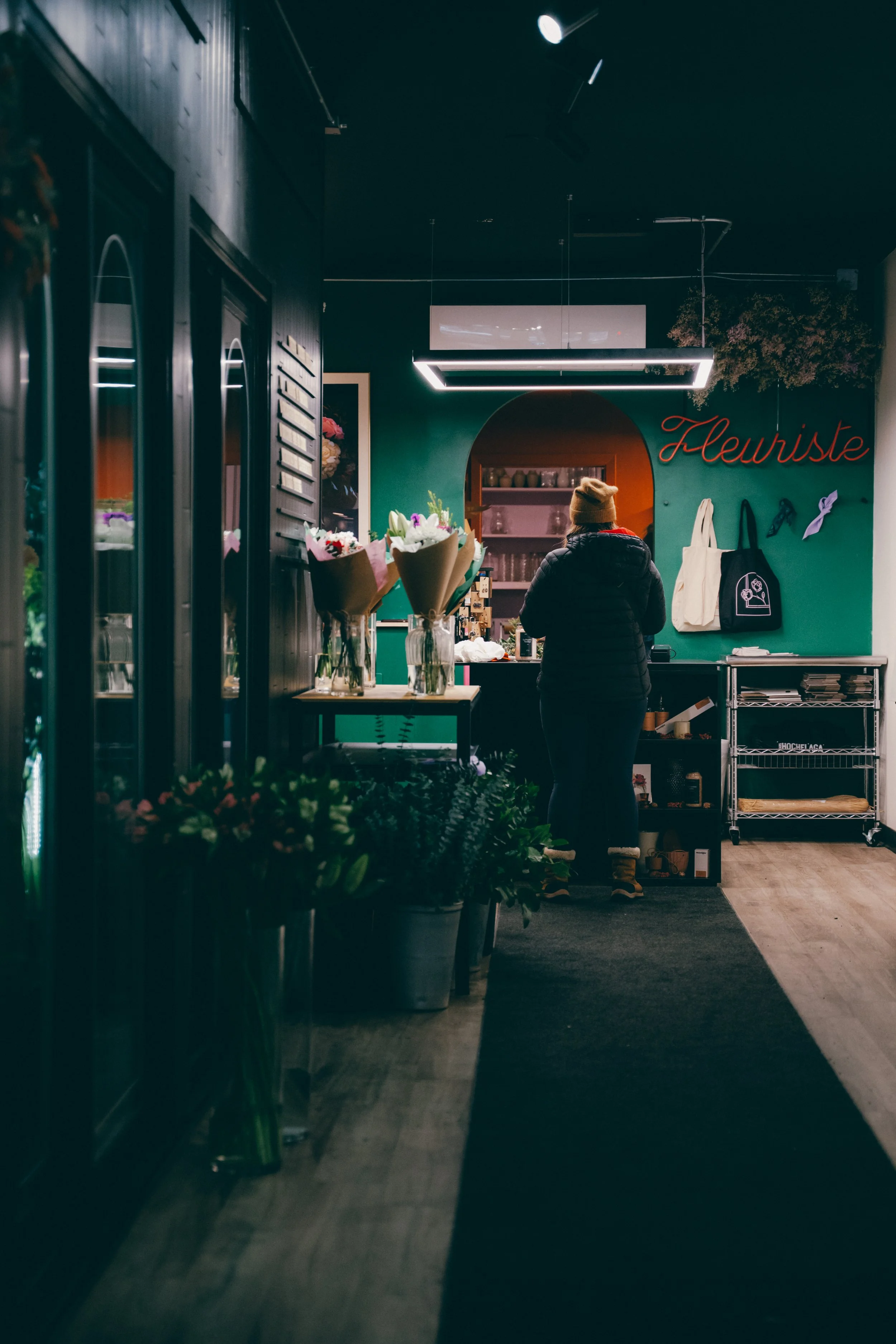 Intérieur d'une boutique de fleurs avec une personne vue de dos qui porte un bonnet marron, urne des bouquets de fleurs dans des vases, un mur vert avec le mot 'Fleuriste' en rouge, sacs en toile, multitude de plantes en pots, et une étagère en métal