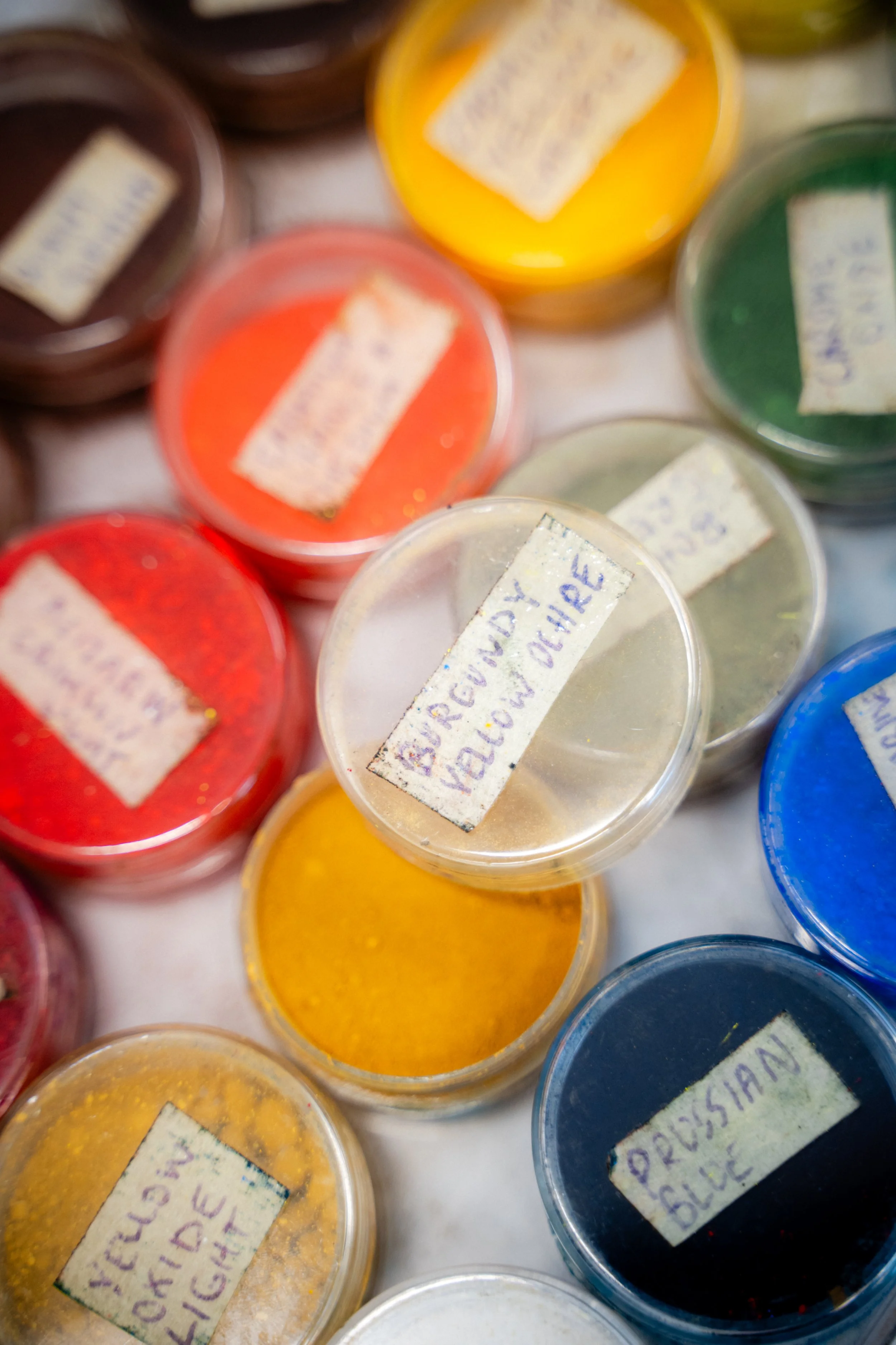 Petits pots de pigments de différentes couleurs, avec des étiquettes manuscrites indiquant les couleurs, comme rouge, jaune, vert, bleu, et d'autres couleurs.
