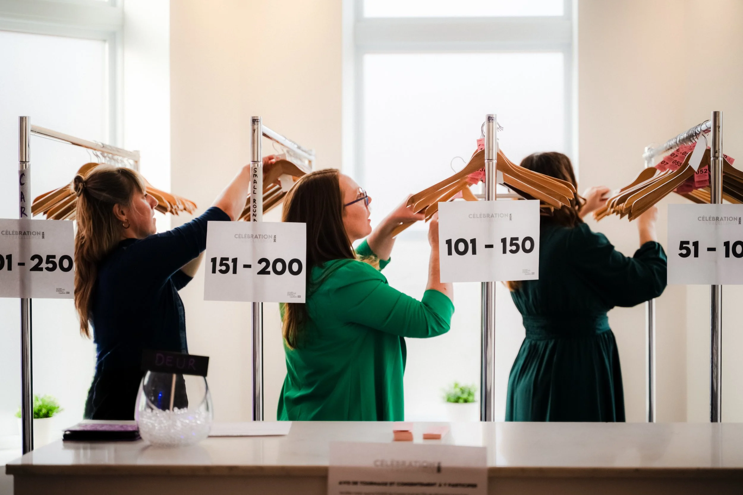 Trois femmes organisent les cintres sur des portants dans le vestiaire d'une salle de réception. Il y a une table avec un verre d'eau et un pot de plantes vertes en arrière-plan.