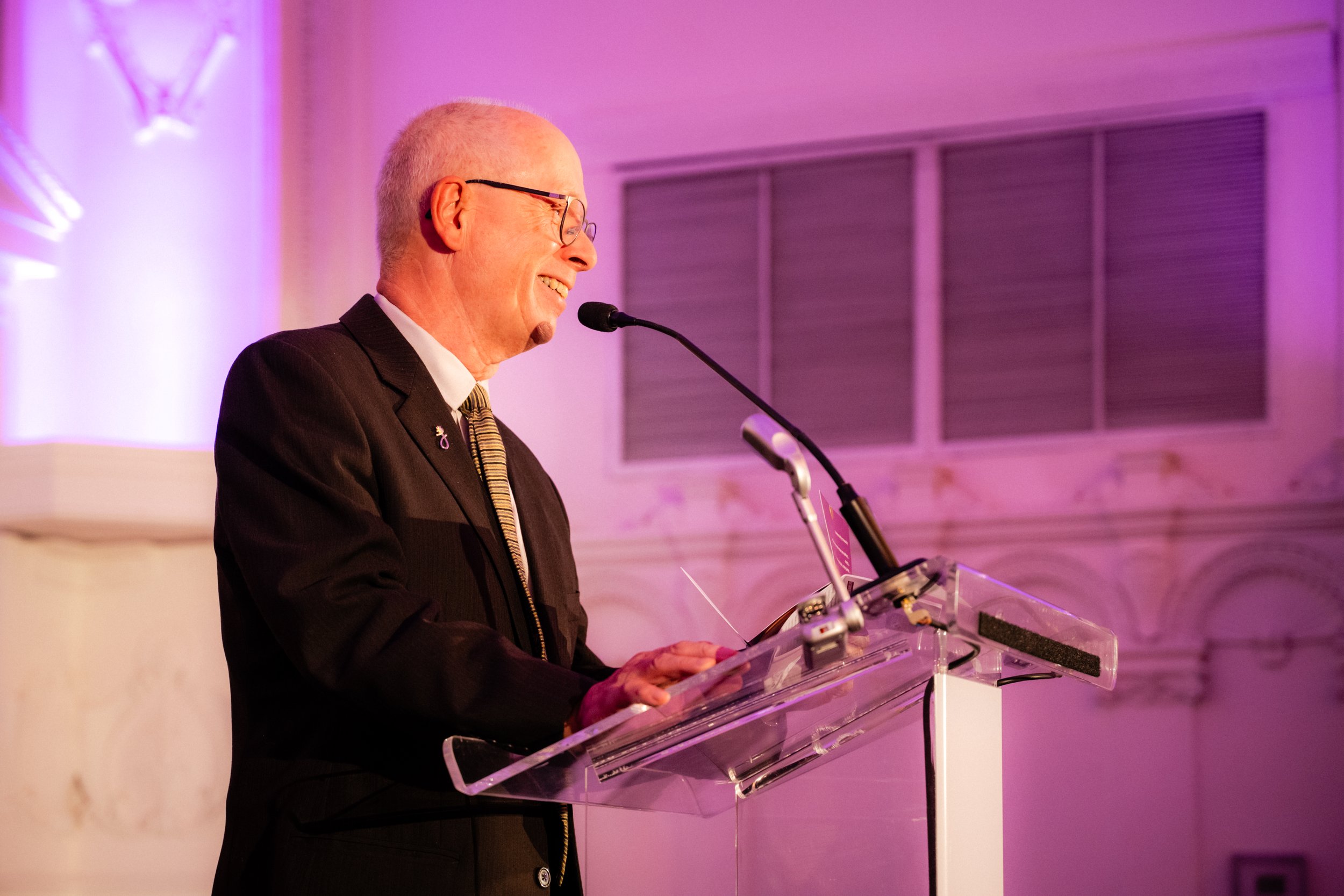 Un homme âgé avec des lunettes parle lors d'une conférence sous un éclairage violet, derrière un pupitre en verre.