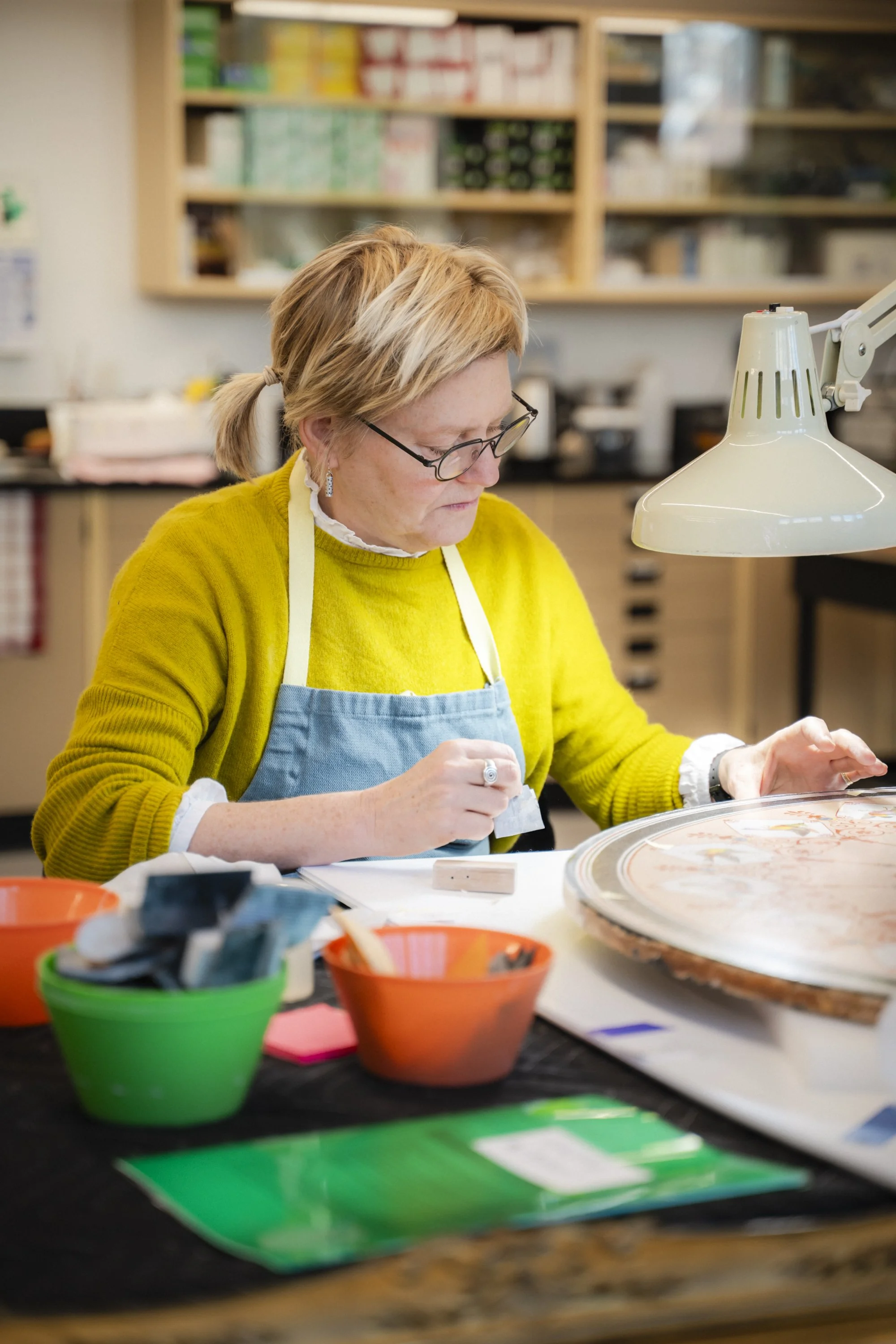 Une femme portant des lunettes, un pull jaune et un tablier bleu, travaille sur un projet créatif dans un atelier, entourée de boîtes de peinture colorées et de matériel artistique.