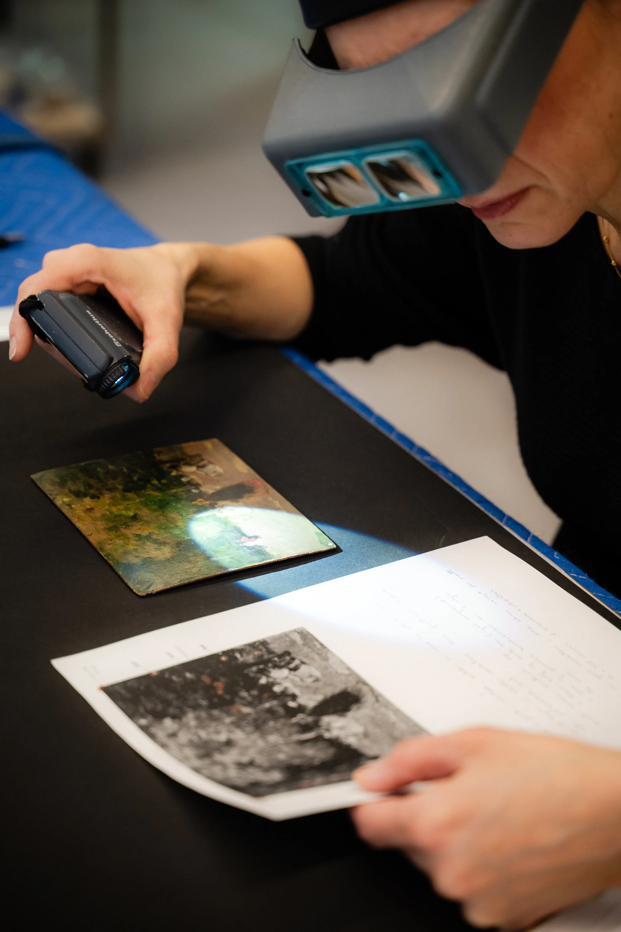 Une personne portant des loupes professionnelles travaille sur une photo ancienne sur une table noire, avec des notes manuscrites et des images de l'œuvre originale imprimées autour.