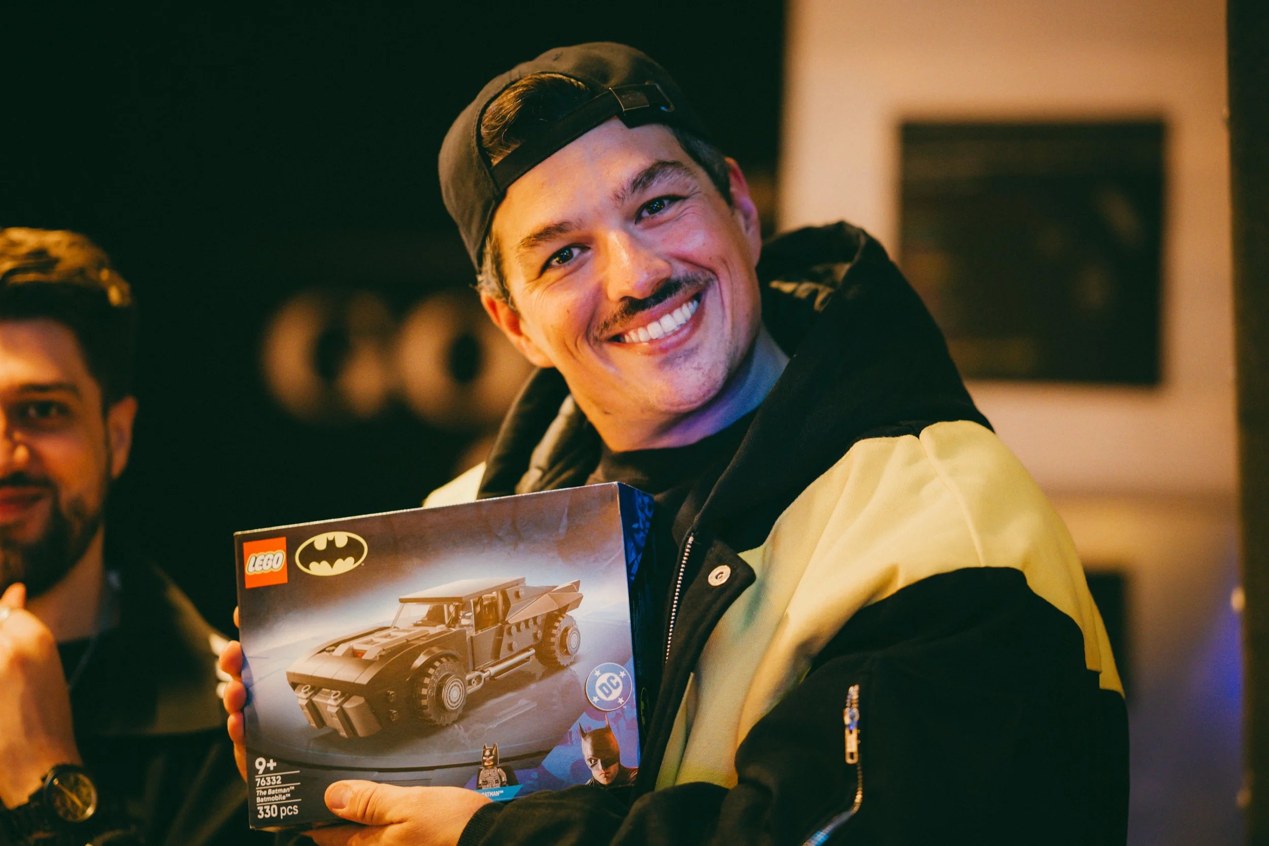 Un jeune homme souriant, portant une casquette sombre à l'envers, tient un coffret LEGO Batman avec une voiture de Batman dessus. Il semble heureux d'avoir reçu le jouet.