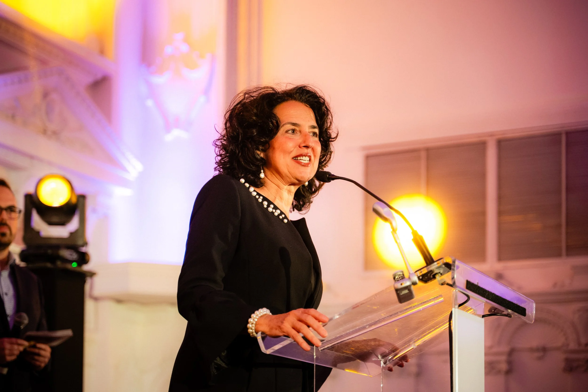 Une femme parlant lors d'un discours ou d'une présentation, derrière un pupitre transparent, dans une salle ornée de lumières chaudes et décorations élégantes.