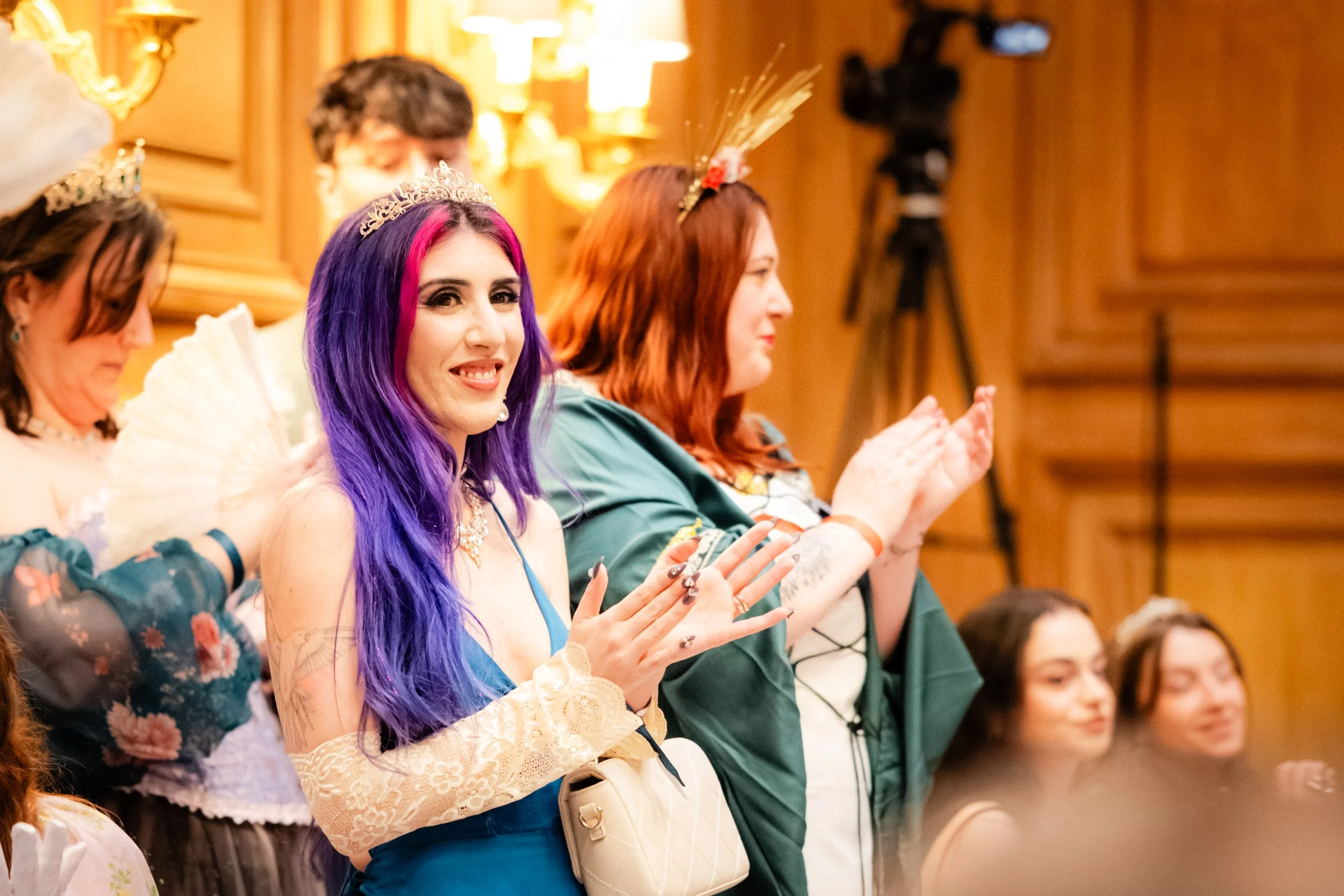 Des femmes portant des couronnes, applaudissant lors d'une cérémonie ou d'un événement. La femme au centre a des cheveux violets et roses et un sourire visible.