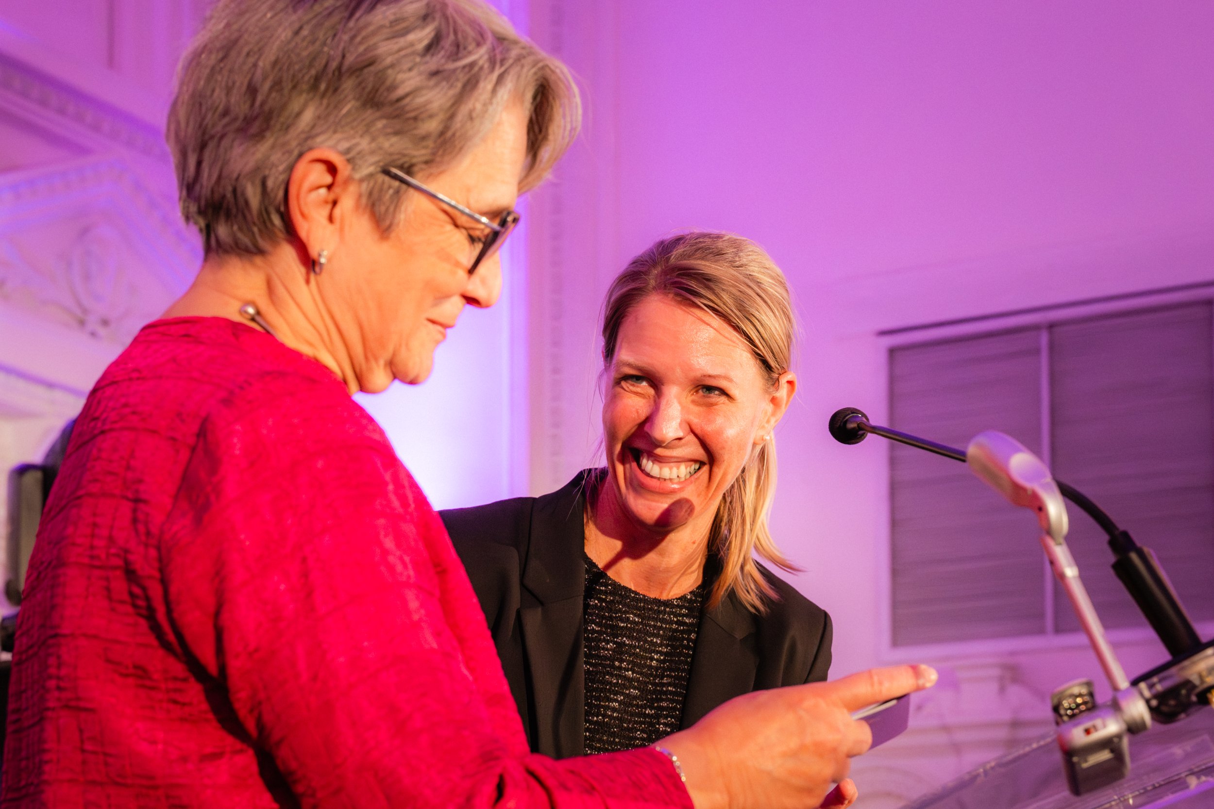 Deux femmes souriantes discutent sur scène avec un micro, face à un fond violet, lors d'une présentation ou d'un événement.