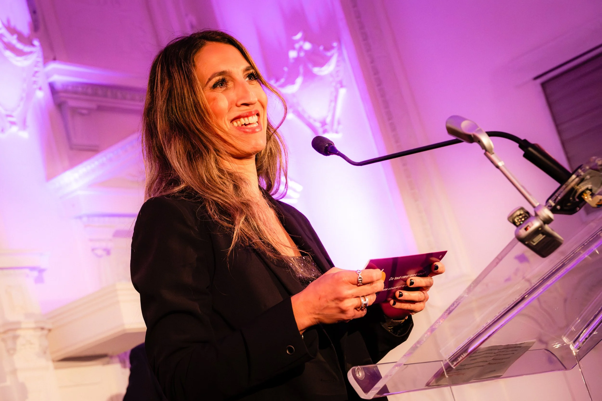 Une femme souriante donne un discours derrière un pupitre en verre dans une salle élégante avec éclairage violet.
