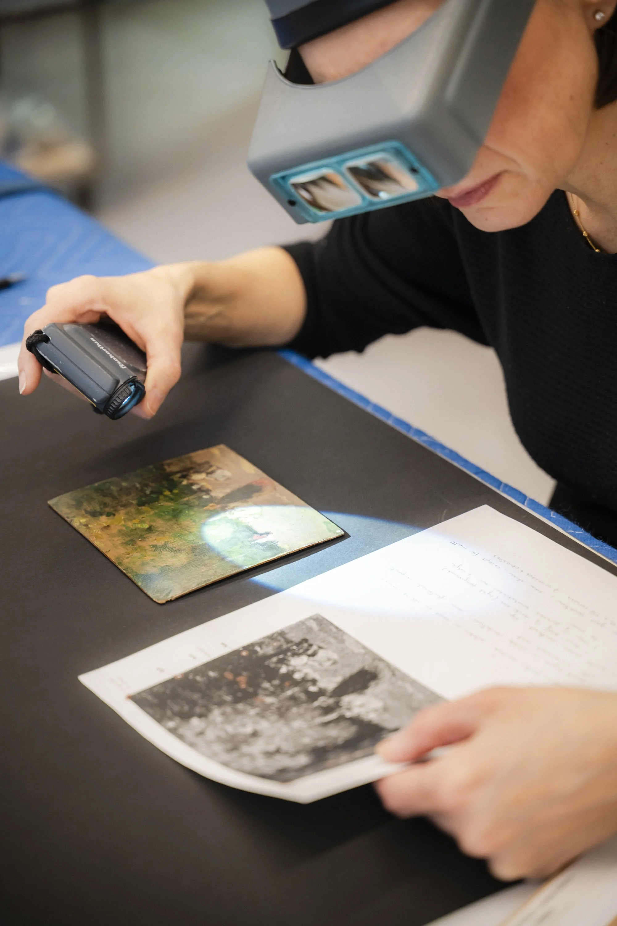 Une personne portant des loupes professionnelles travaille sur une photo ancienne sur une table noire, avec des notes manuscrites et des images de l'œuvre originale imprimées autour.