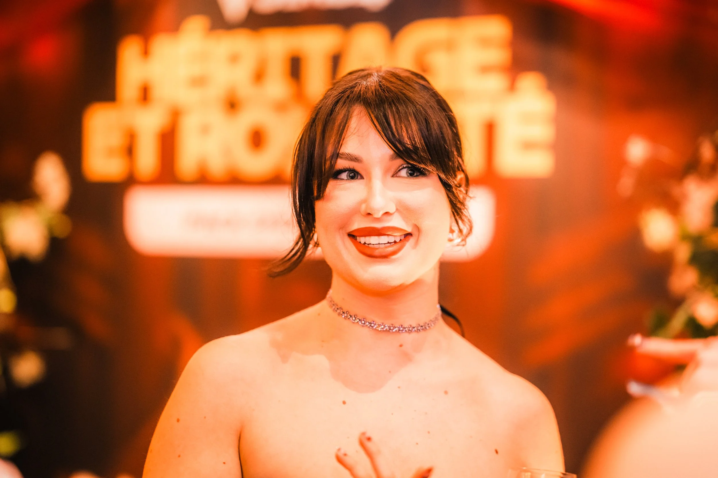 Une jeune femme souriante avec des cheveux bruns foncés et une coiffure en boucles, portant un collier en chaîne, pose à proximité d'une lumière orange et d'un fond flou où l'on peut lire partiellement le mot "HEAVEN" en lettres lumineuses, dans une 