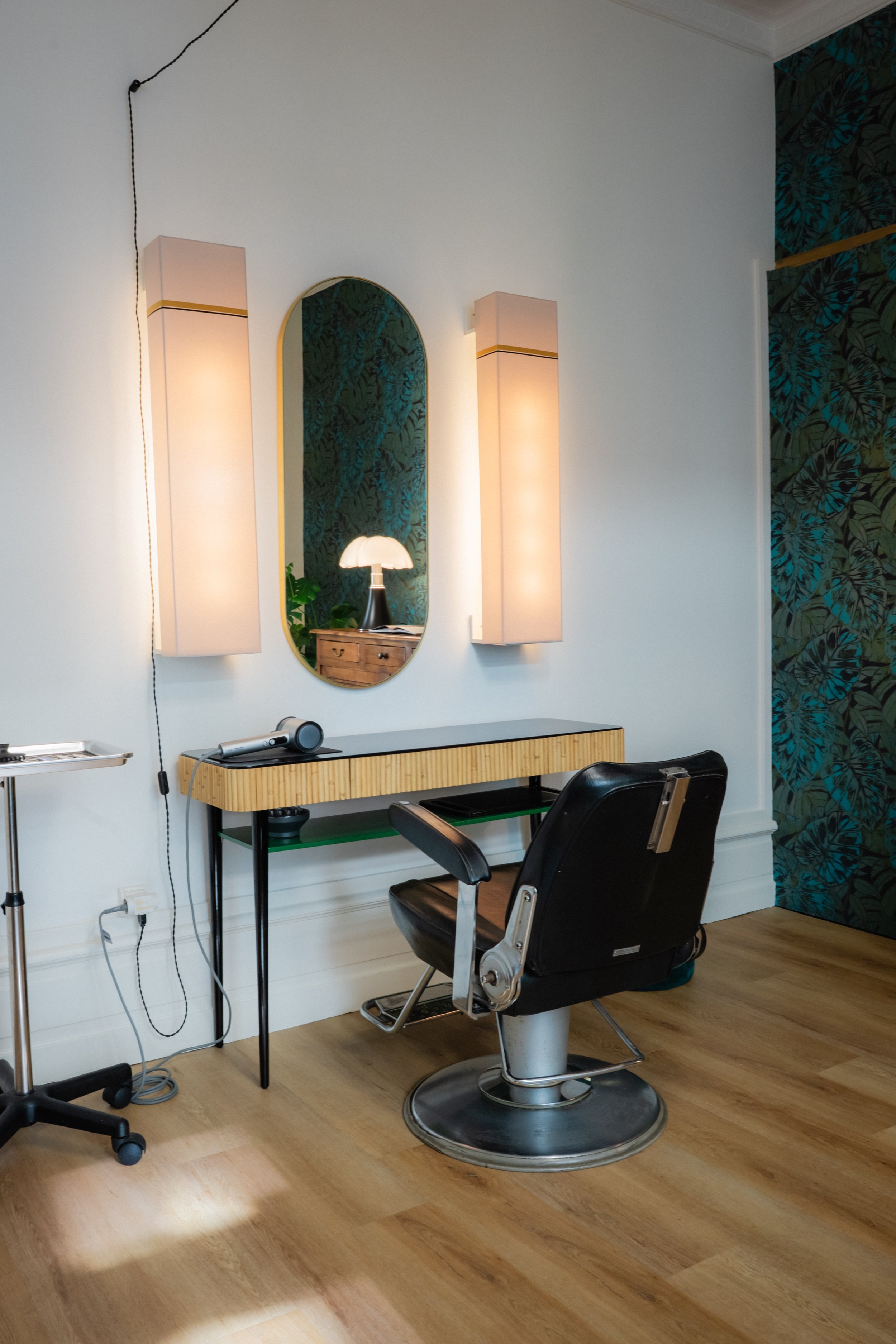 Un salon de coiffure avec un fauteuil de coiffure en cuir noir, un miroir ovale, deux appliques murales, un bureau en bois clair design avec un sèche-cheveux, un siège en cuir noir, et un mur avec un motif végétal