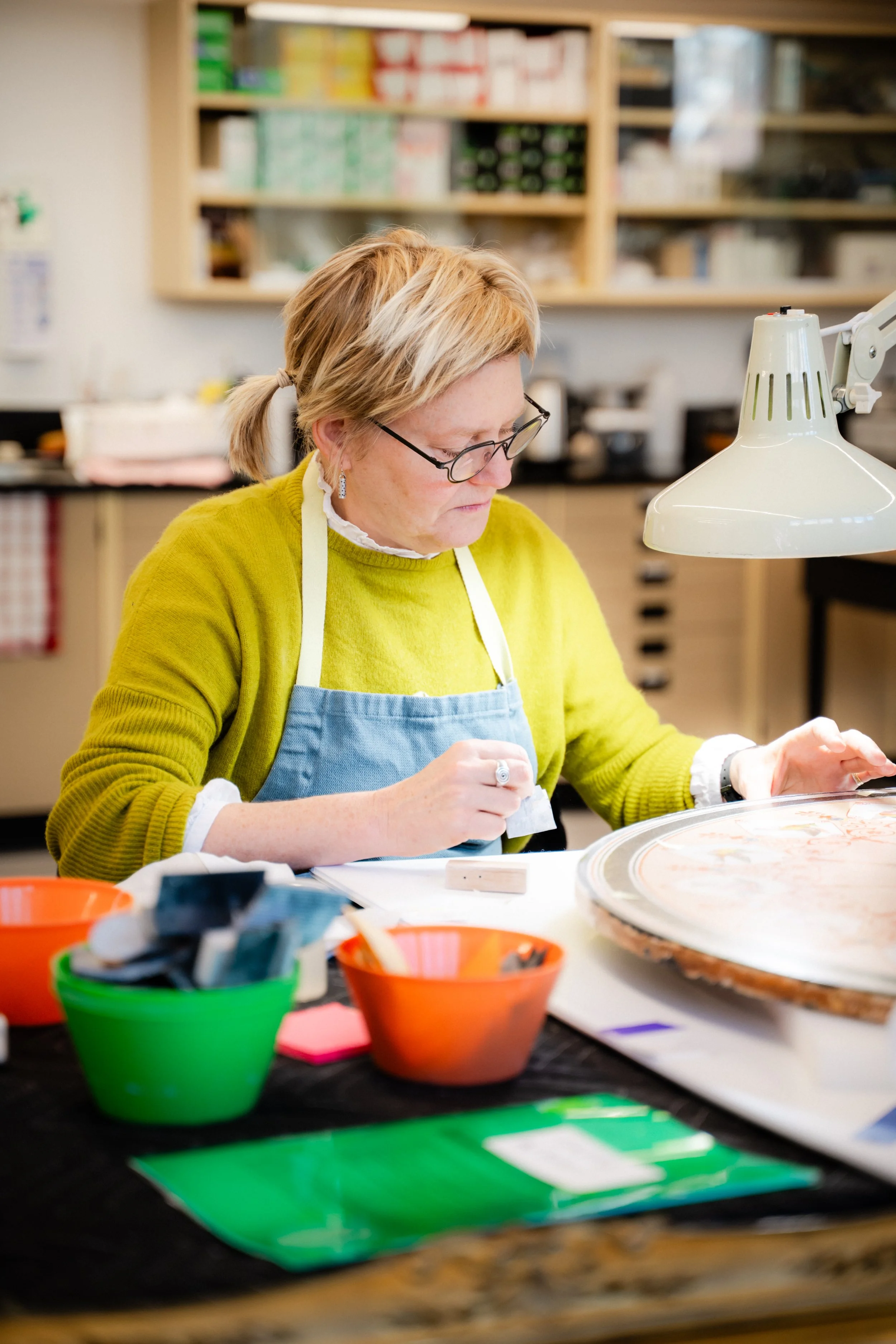 Une femme portant des lunettes, un pull jaune et un tablier bleu, travaille sur un projet créatif dans un atelier, entourée de boîtes de peinture colorées et de matériel artistique.