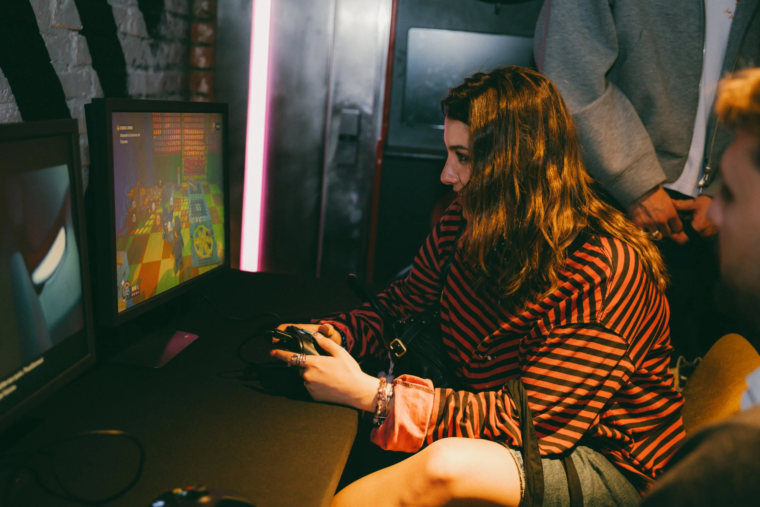 Une jeune femme avec des cheveux ondulés bruns joue à un jeu vidéo dans une salle de jeu, assise devant un écran d'ordinateur, entourée d'autres personnes.
