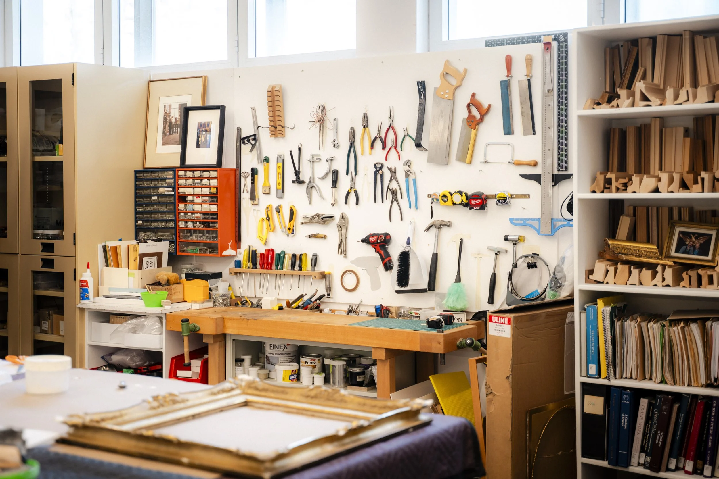 Un atelier ou un espace de bricolage avec un mur où sont suspendus divers outils comme des marteaux, des ciseaux, des pinces, des scies, des tournevis et d'autres équipements. Il y a également des étagères avec des livres et des boîtes de rangement, 
