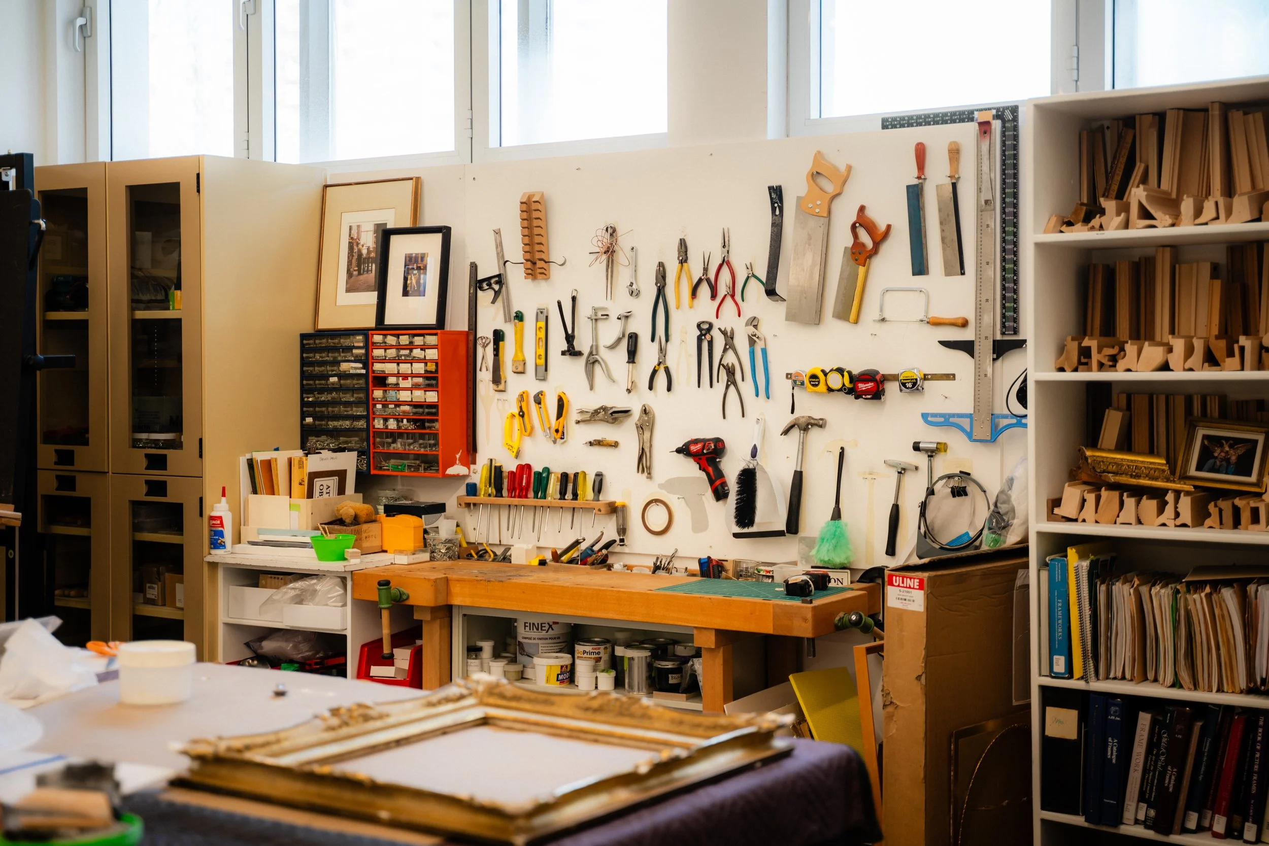 Un atelier ou un espace de bricolage avec un mur où sont suspendus divers outils comme des marteaux, des ciseaux, des pinces, des scies, des tournevis et d'autres équipements. Il y a également des étagères avec des livres et des boîtes de rangement, 
