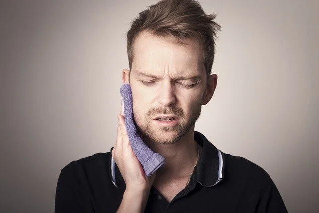 Un homme avec une expression de douleur, tenant une compresse glacée sur sa joue, suggérant qu'il a mal à la mâchoire ou au visage.