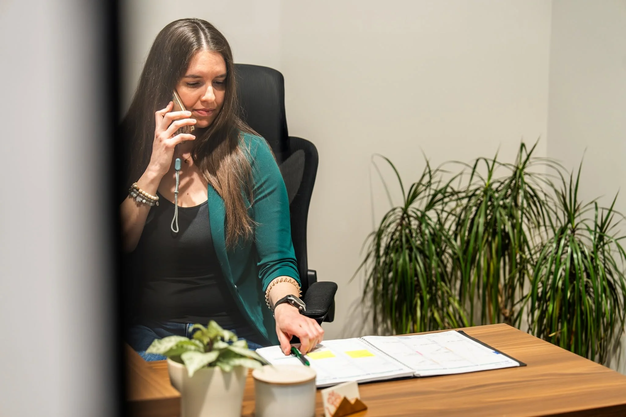 Une femme assise à une table avec un agenda ouvert, parlant au téléphone, dans un environnement de bureau moderne avec un mur blanc, une plante verte et une décoration sur la table.