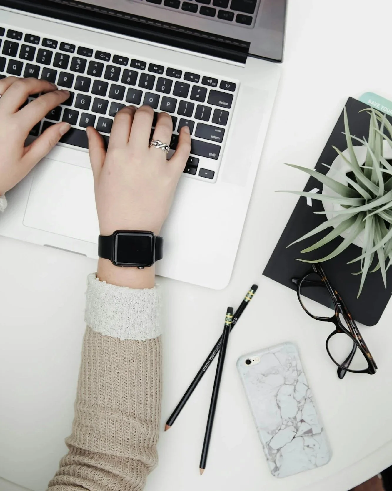 Vue du dessus d'un bureau avec une personne tapant sur un ordinateur portable, des lunettes, deux stylos, un téléphone portable avec une coque en marbre, un carnet noir avec une plante en pot et un crayon, tous disposés sur un bureau blanc.