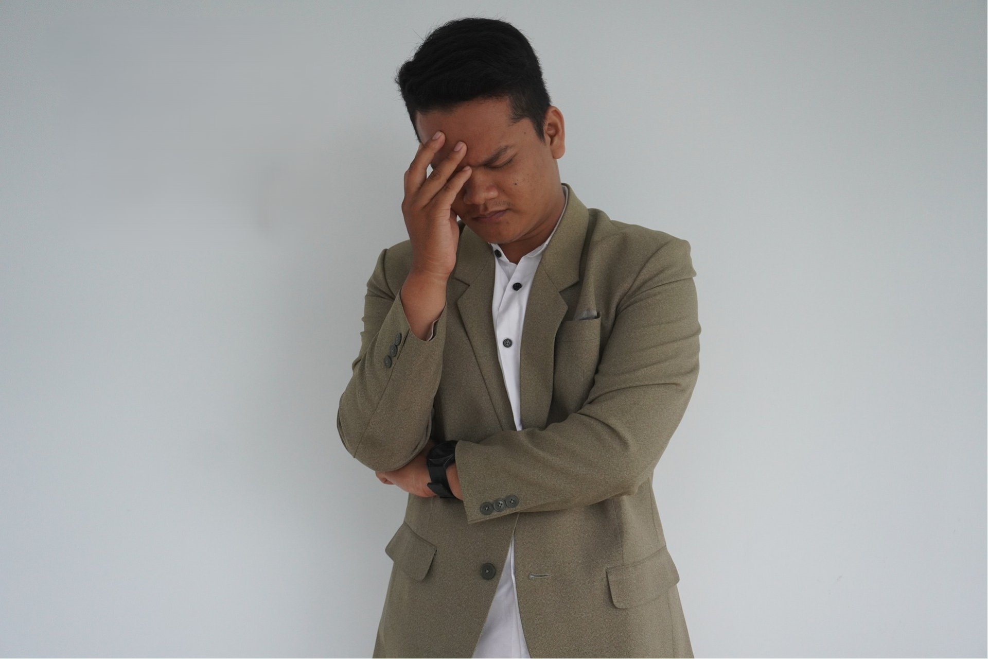 Un homme habillé en costume beige, se tient contre un mur blanc avec une expression de frustration ou de réflexion, tenant sa tête avec sa main.