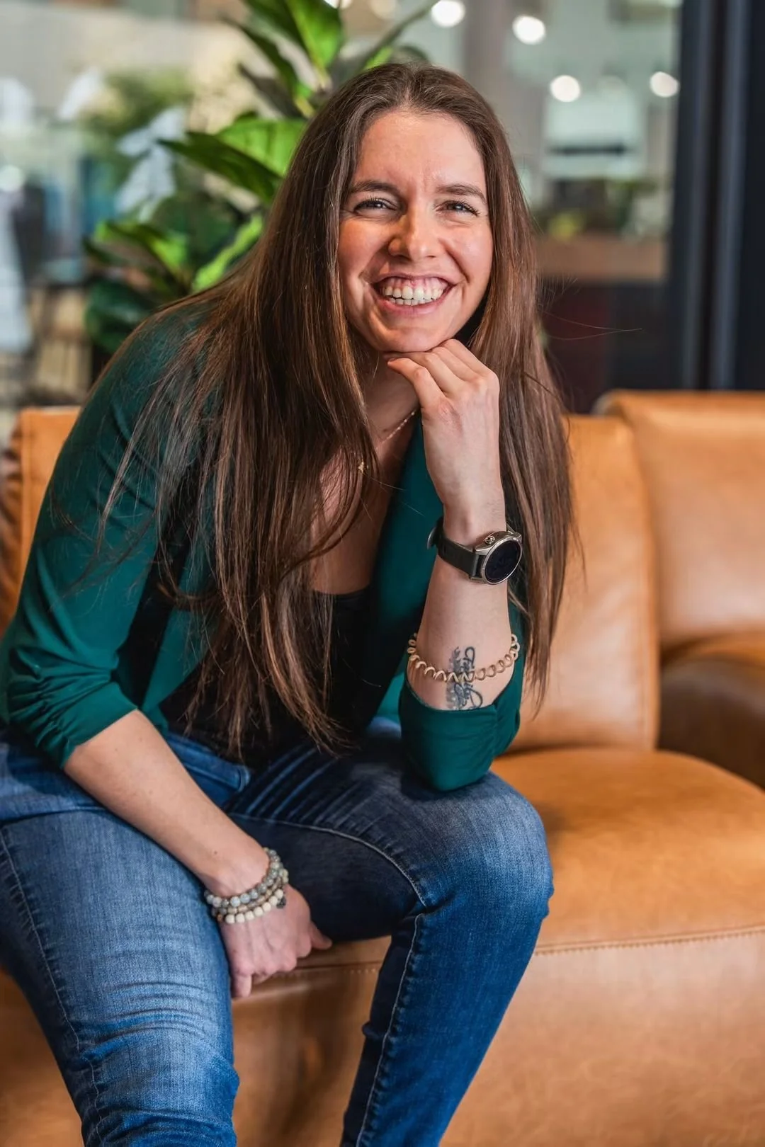 Une femme avec de longs cheveux bruns souriant, assise sur un canapé en cuir orange, portant un bracelet, une montre, une chemise verte à manches longues et un jean, dans un intérieur moderne avec un grand plante verte en arrière-plan.