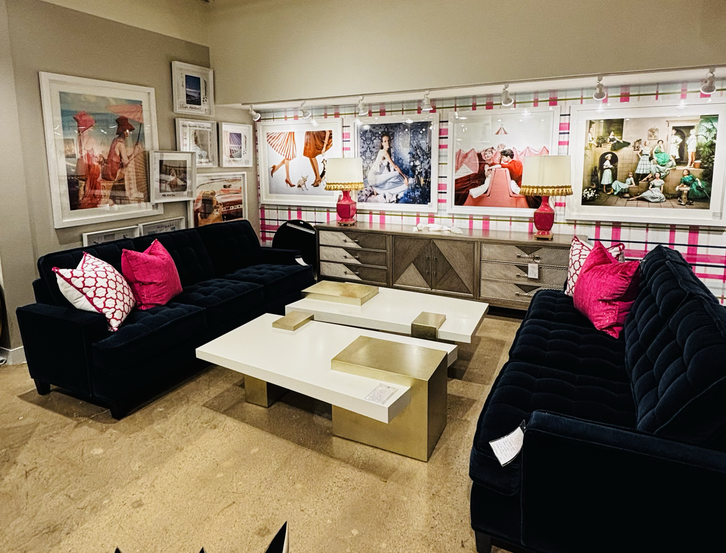 Living room with black sofas with pink and white patterned pillows, a white coffee table with brass accents, a wood sideboard, and a wall decorated with framed artwork, including fashion photographs.