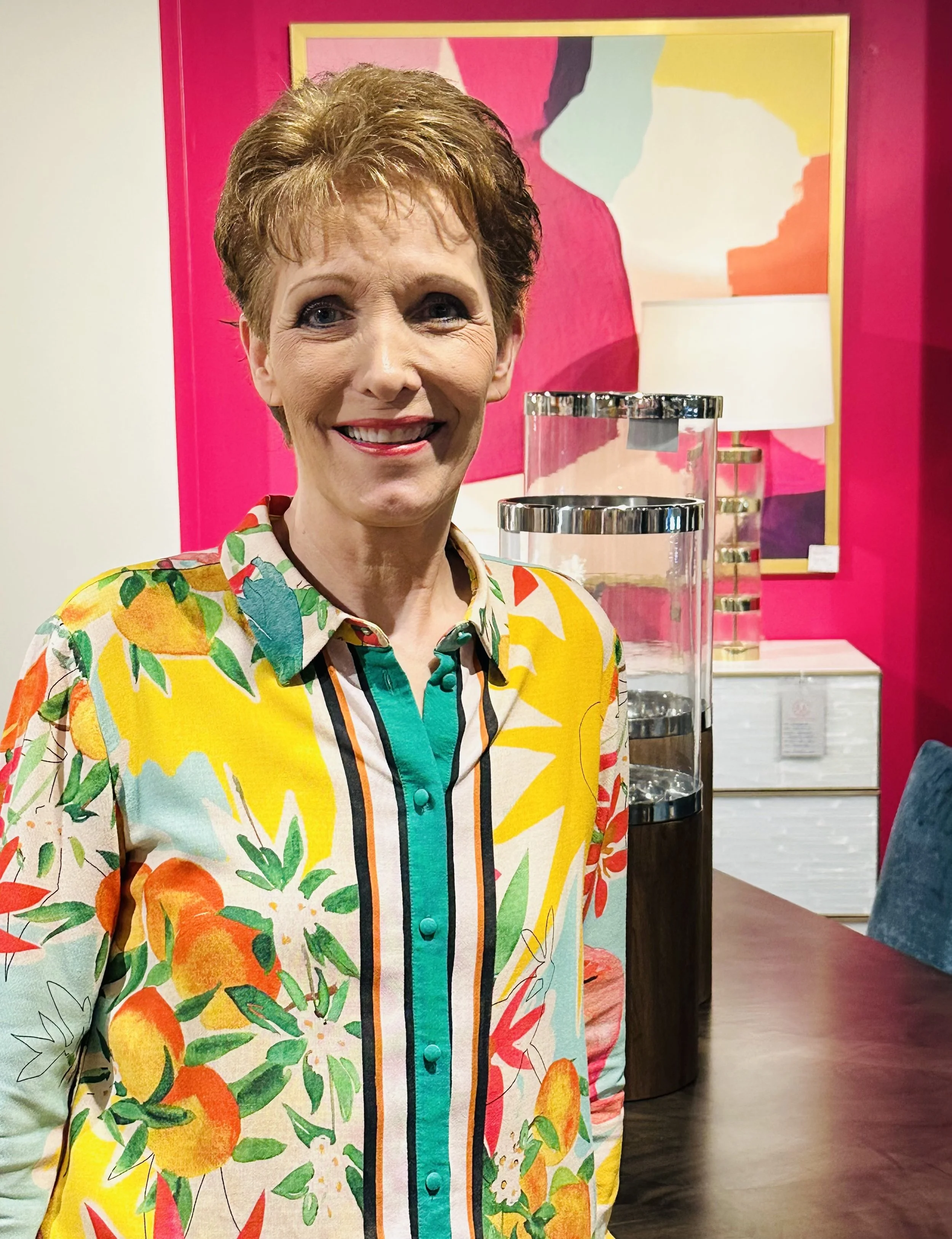 Middle-aged woman with short reddish-brown hair, smiling, wearing a colorful floral blouse with orange, yellow, green, and blue patterns. She is standing in front of a pink wall with abstract artwork and a modern glass water dispenser.