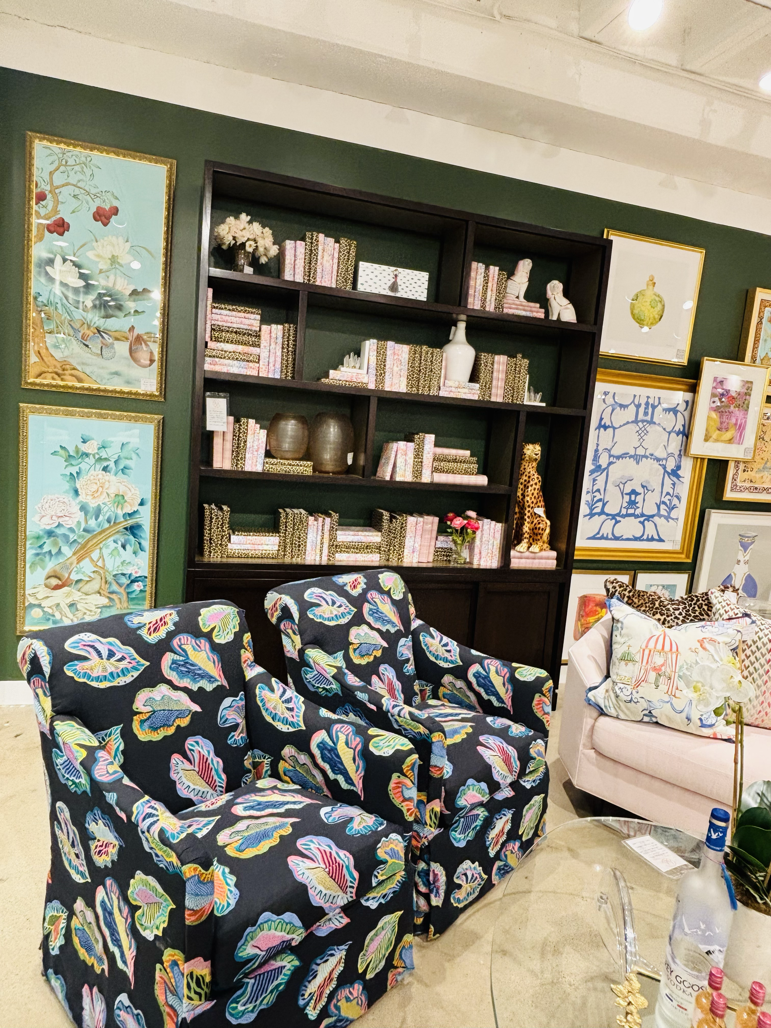 Interior of a room with green walls decorated with framed artwork. There are shelves filled with books and decorative objects, including a white vase and small animal statues. In front, a colorful armchair with a floral pattern and a pink sofa with pillows are visible. On the side, a table with bottles and flowers.