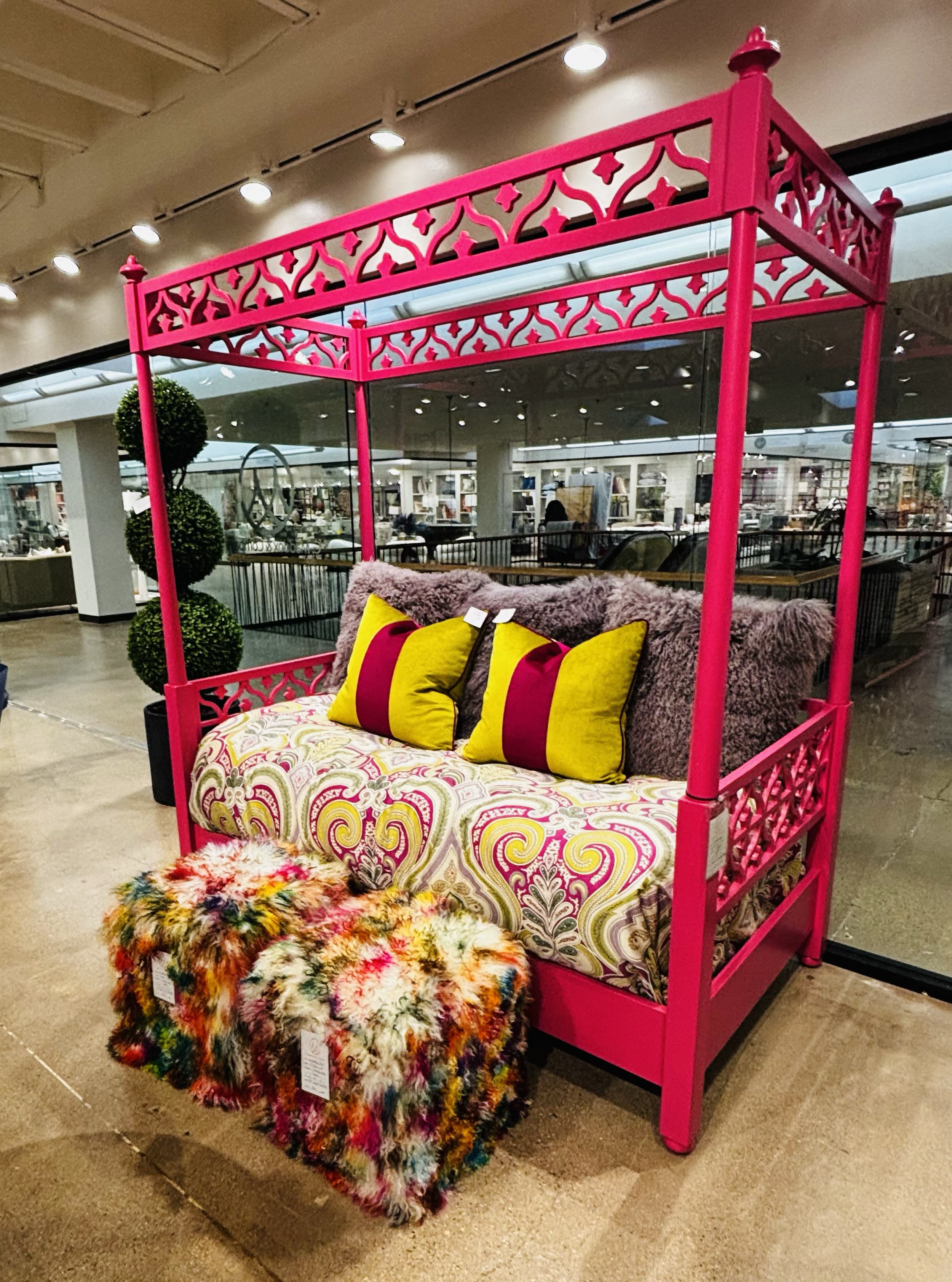 A vibrant pink painted bed with ornate cutout decorations, decorated with colorful cushions, a fuzzy throw, and a multicolored faux fur rug at the foot, displayed in a store.