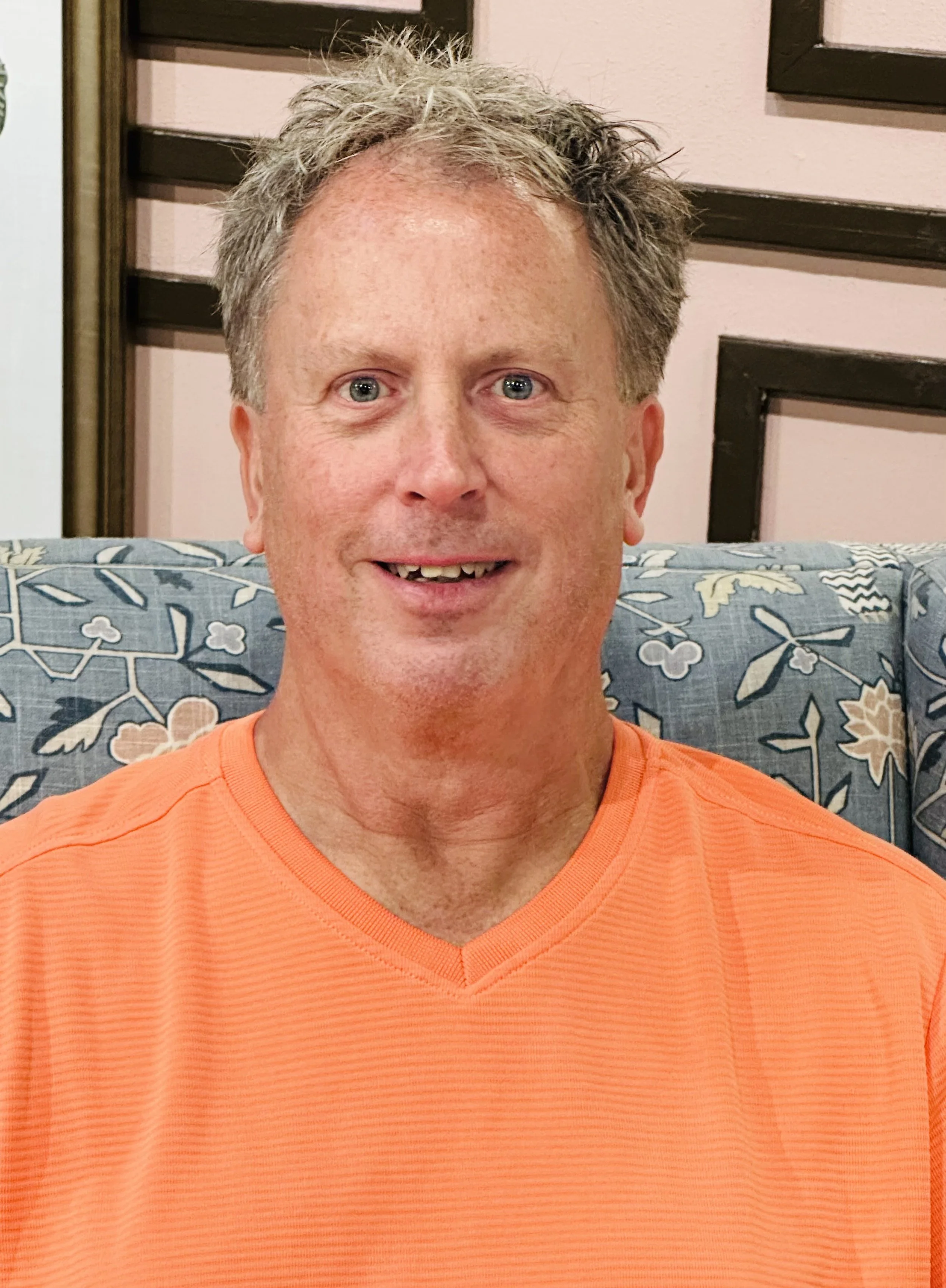 A middle-aged man with short, curly gray hair, blue eyes, and light skin, smiling while sitting in a room with pink walls, black framed wall art, and a patterned blue cushion, wearing an orange athletic shirt.