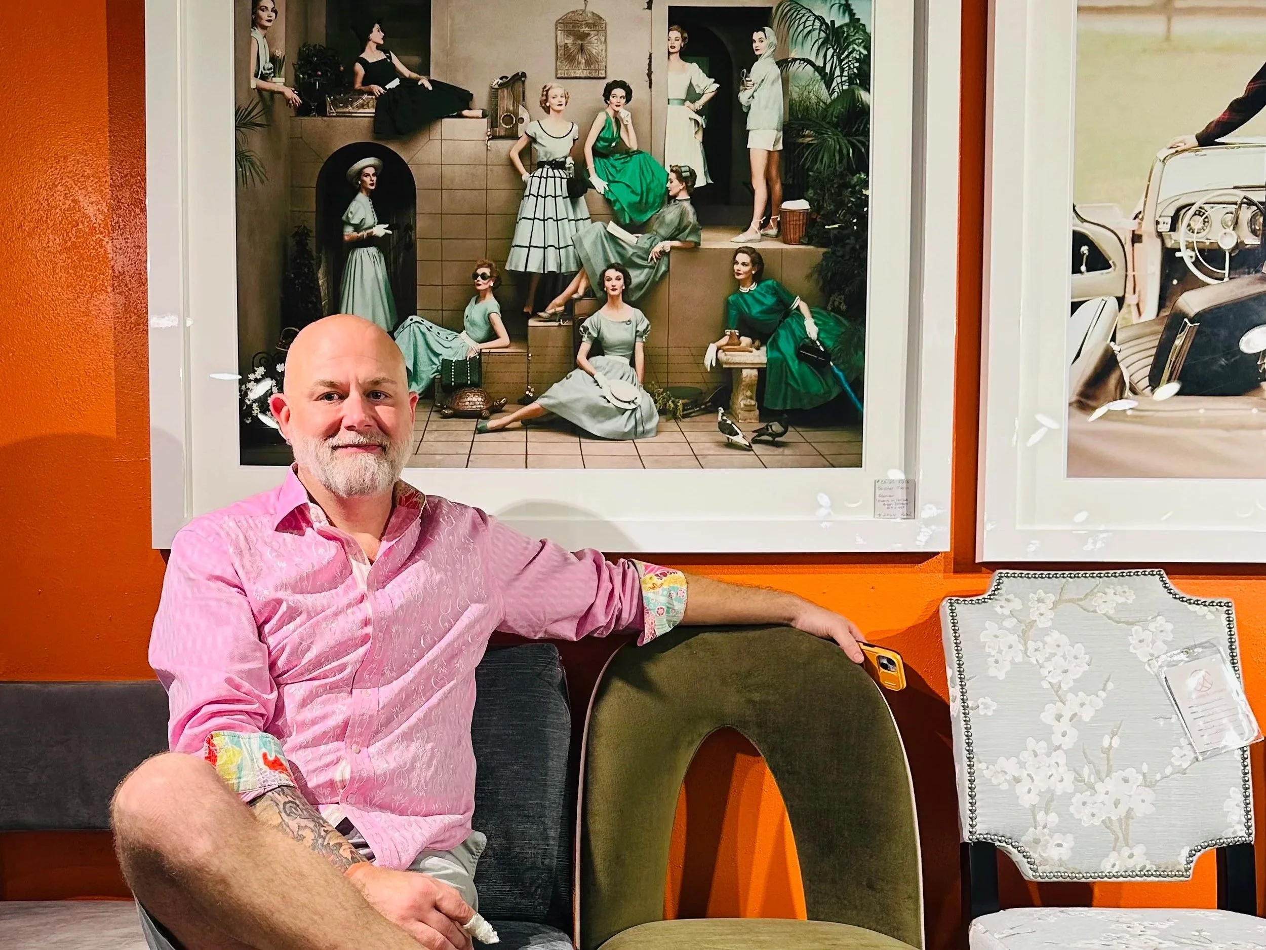 A man with a beard and tattoos wearing a pink shirt sits in front of a colorful artwork featuring mannequins in vintage clothing, with the man holding a yellow phone and resting his arm on a chair.